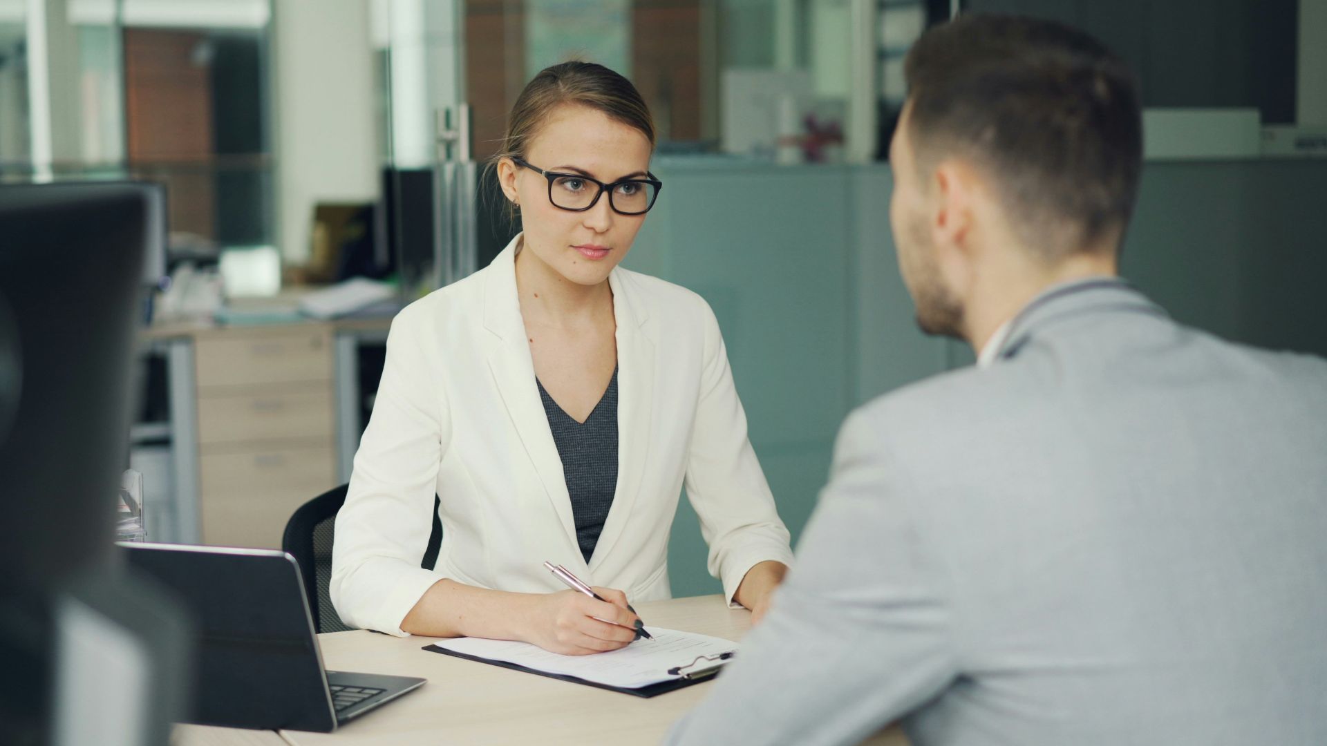 Woman in glasses interviews man at office desk.
