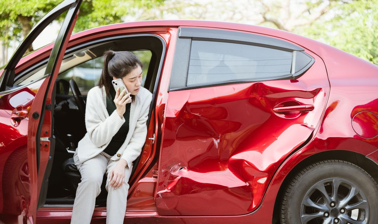 Car accident crash, Cars insurance protection, Claim collision, Auto emergency. A woman is sitting in a red car with a smashed front end. She is talking on her cell phone.