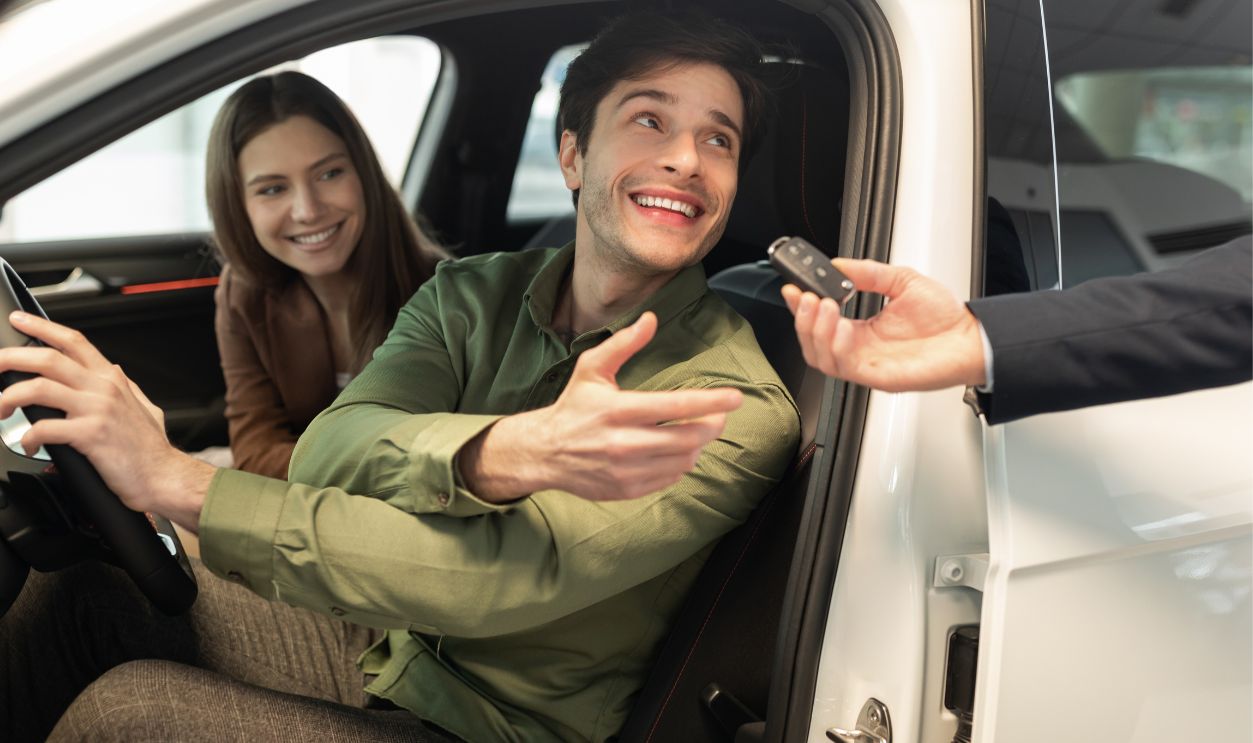 Happy millennial couple taking car key from auto salesman, sitting inside modern automobile at dealership, panorama. Cheery young family buying new vehicle at modern showroom