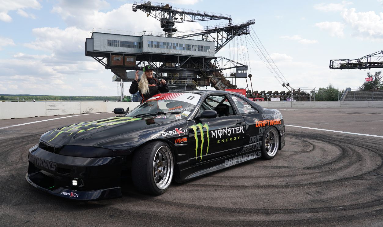 14 August 2019, Saxony-Anhalt, Gräfenhainichen: The driver Tessa Whittock (Great Britain) poses at a Nissan Skyline R33 and the tire tracks of a so-called Donat before the motorsport event Iron Drift King 2019. From 15.-17.08.2019 more than 100 drift drivers from 26 nations drive around the crown of the Iron Drift King 2019 in Ferropolis. 