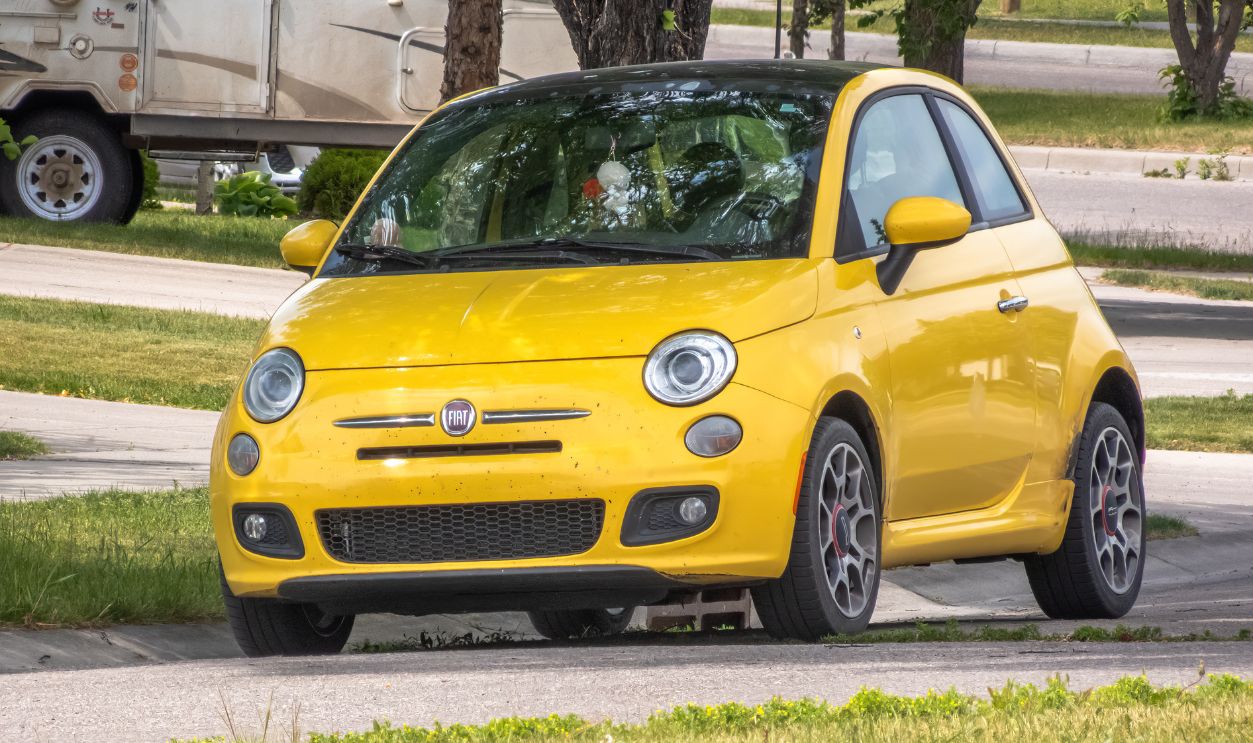  A sunshine yellow Fiat 500 parked on a residential street, a classic car design popular for its fuel efficiency and maneuverability.