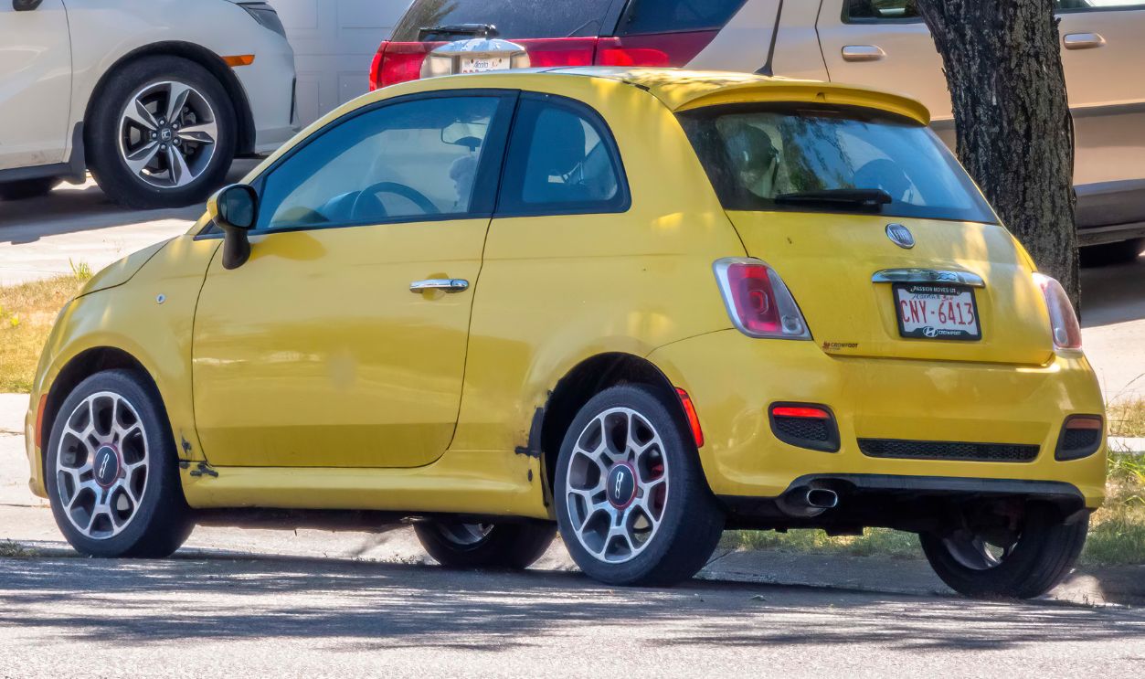 A back view of a sunshine yellow Fiat 500 parked on a residential street, a classic car design popular for its fuel efficiency and maneuverability.