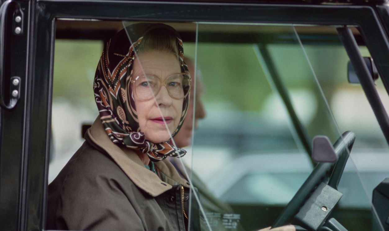 British Royal Queen Elizabeth II, wearing a headscarf and a waxed jacket, driving a Land Rover Defender at the Royal Windsor Horse Show, held at Windsor Home Park in Windsor, Berkshire, England, 12th May 1989.