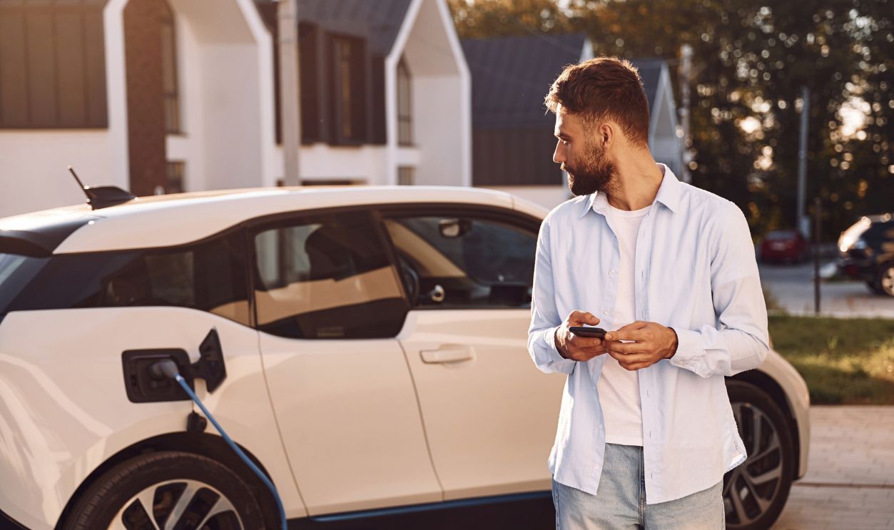 Standing and looking at vehicle that is being charging. Young stylish man is with electric car at daytime.