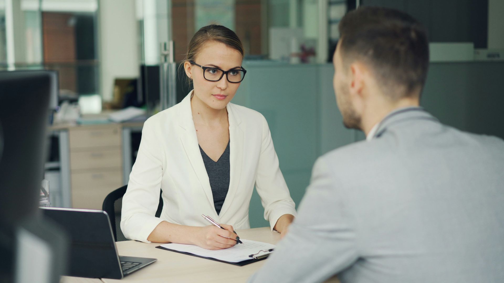 Woman in glasses interviews man at office desk.