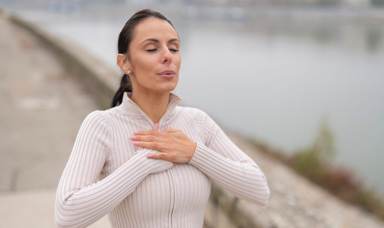 Woman standing outdoors with eyes closed, hands on chest, practicing deep breathing technique
