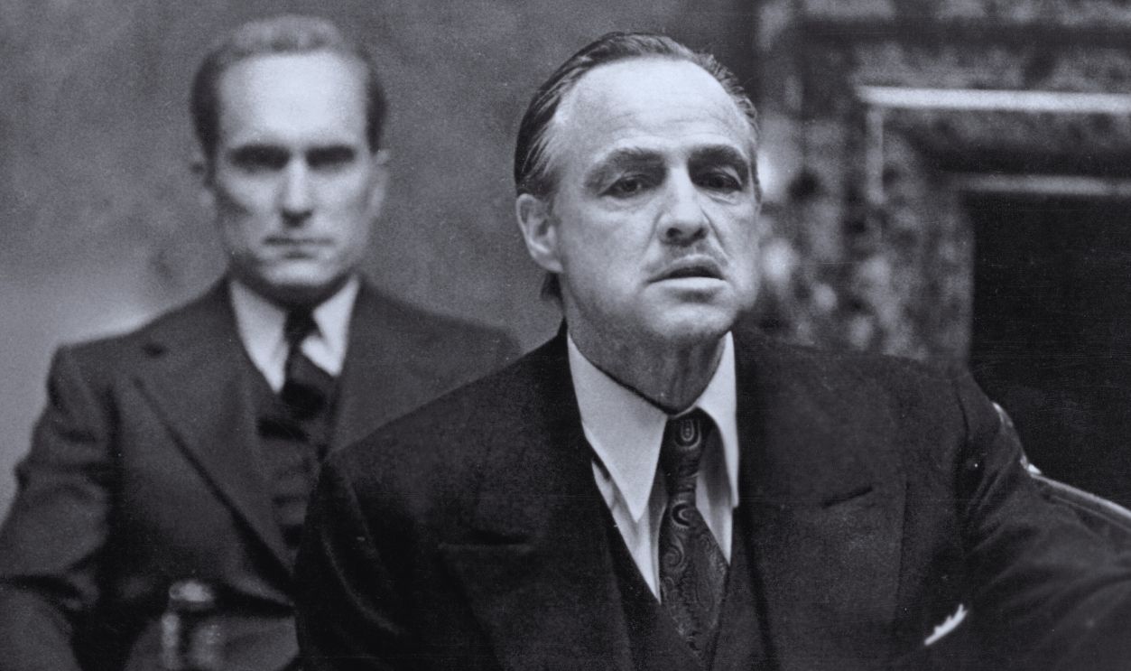 Gettyimages - 57080197, Brando Is 'The Godfather' American actor Marlon Brando (1924 - 2004), in character as mob kingpin Don Vito Corleone, gestures as he sits at a table as colleague and compatriot Robert Duvall, as Tommy Hagen, sits behind him in a scene from the gangster film 'The Godfather,' directed by Francis Ford Coppola, 1972. 