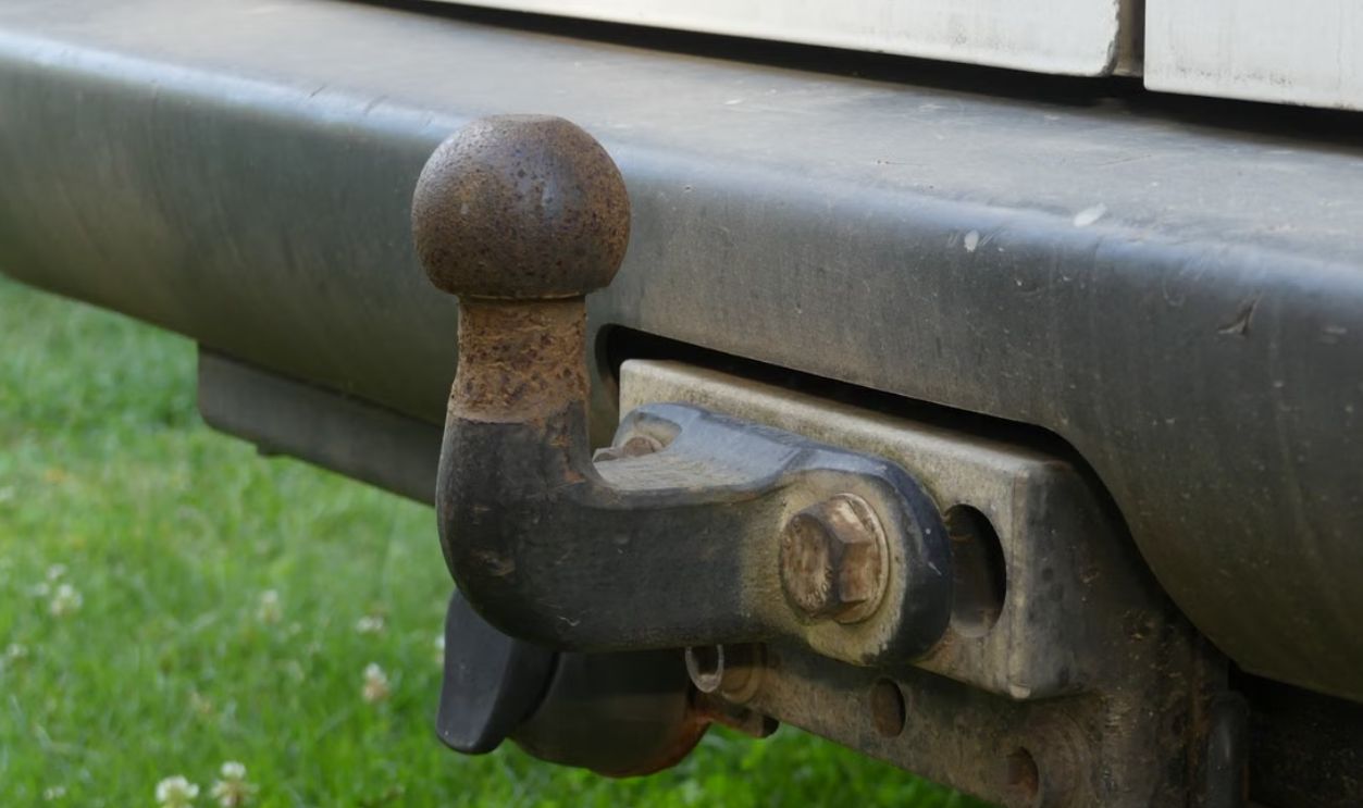 Close-up of a rusty tow hitch on the rear bumper of a vehicle against a grassy background.