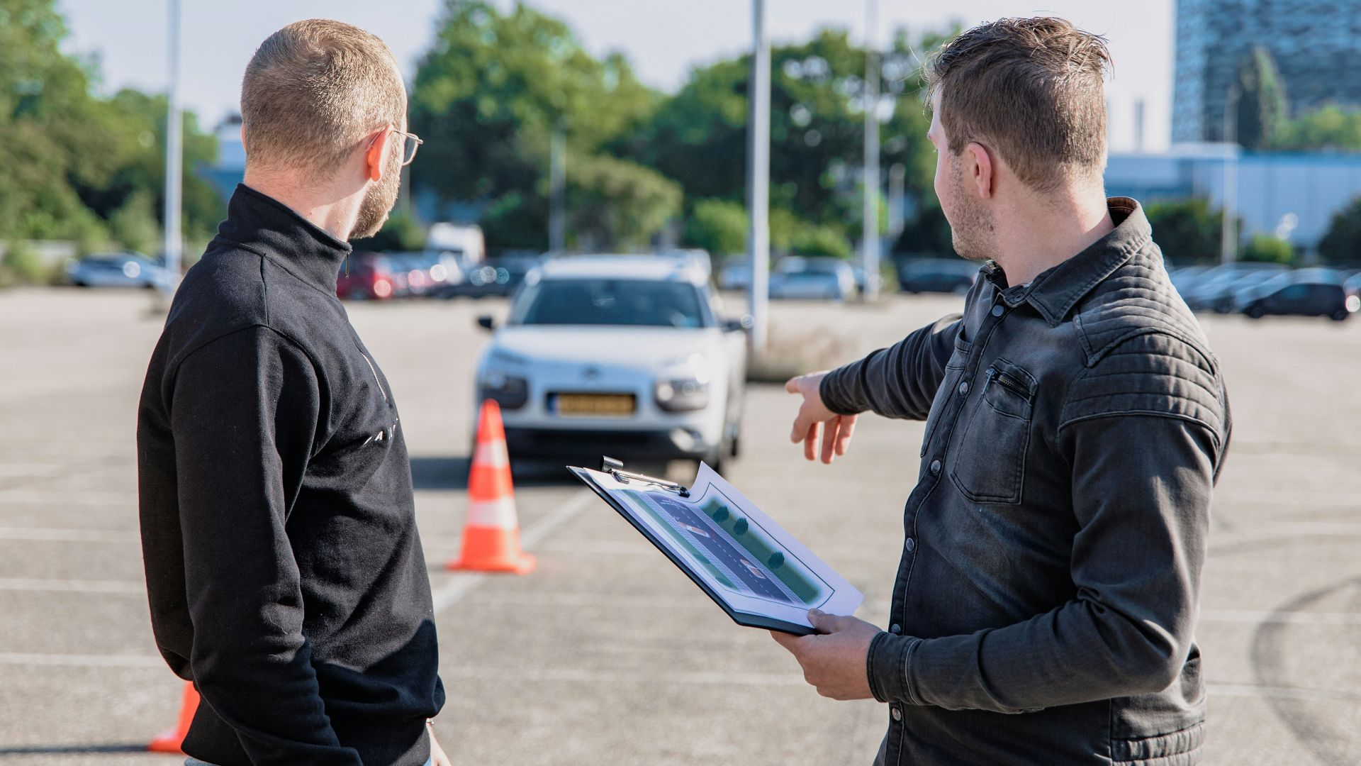 A man pointing toward a car while holding a clipboard for a driving test
