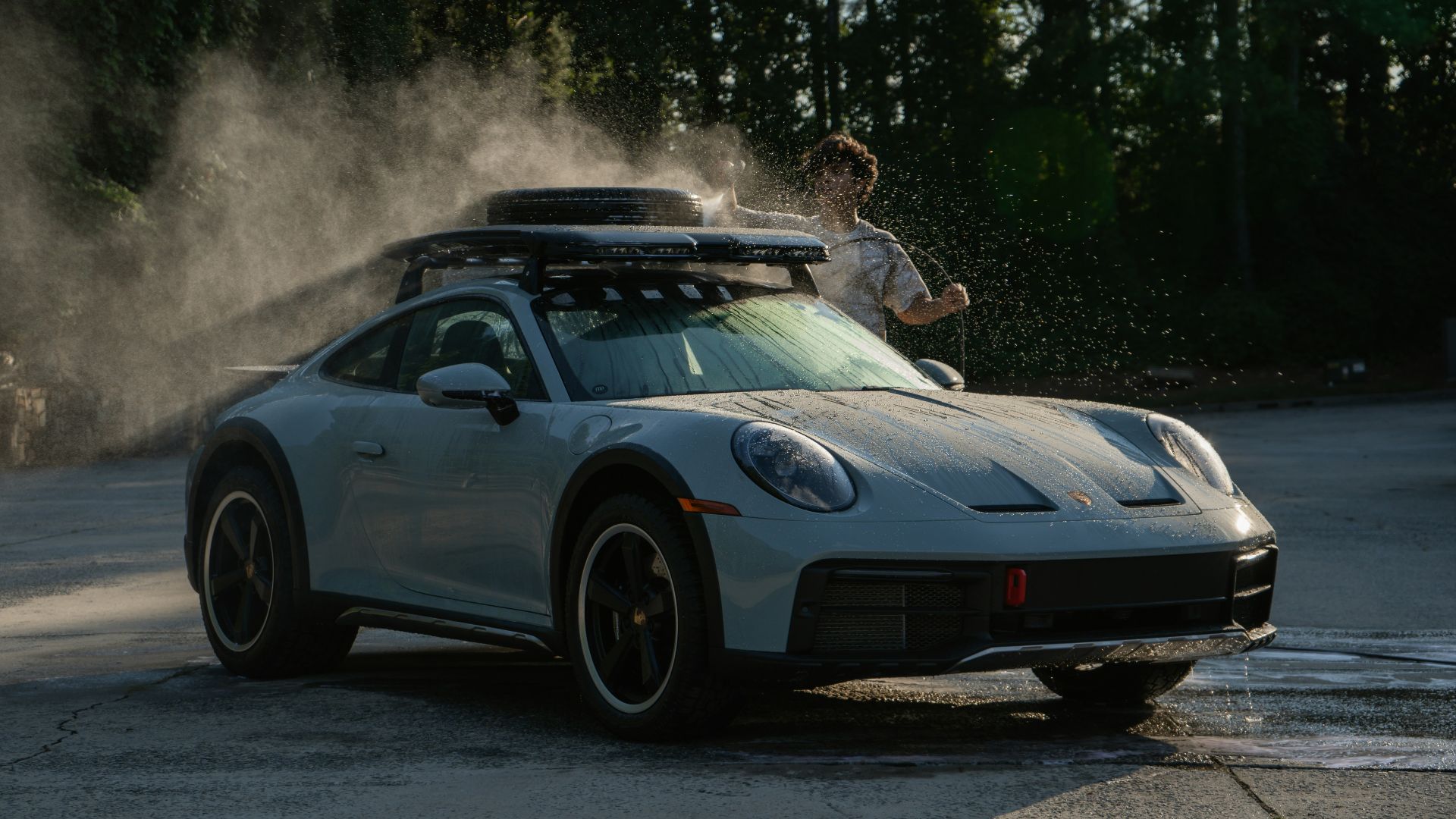 A person washing a light grey sports car with a roof rack outdoors