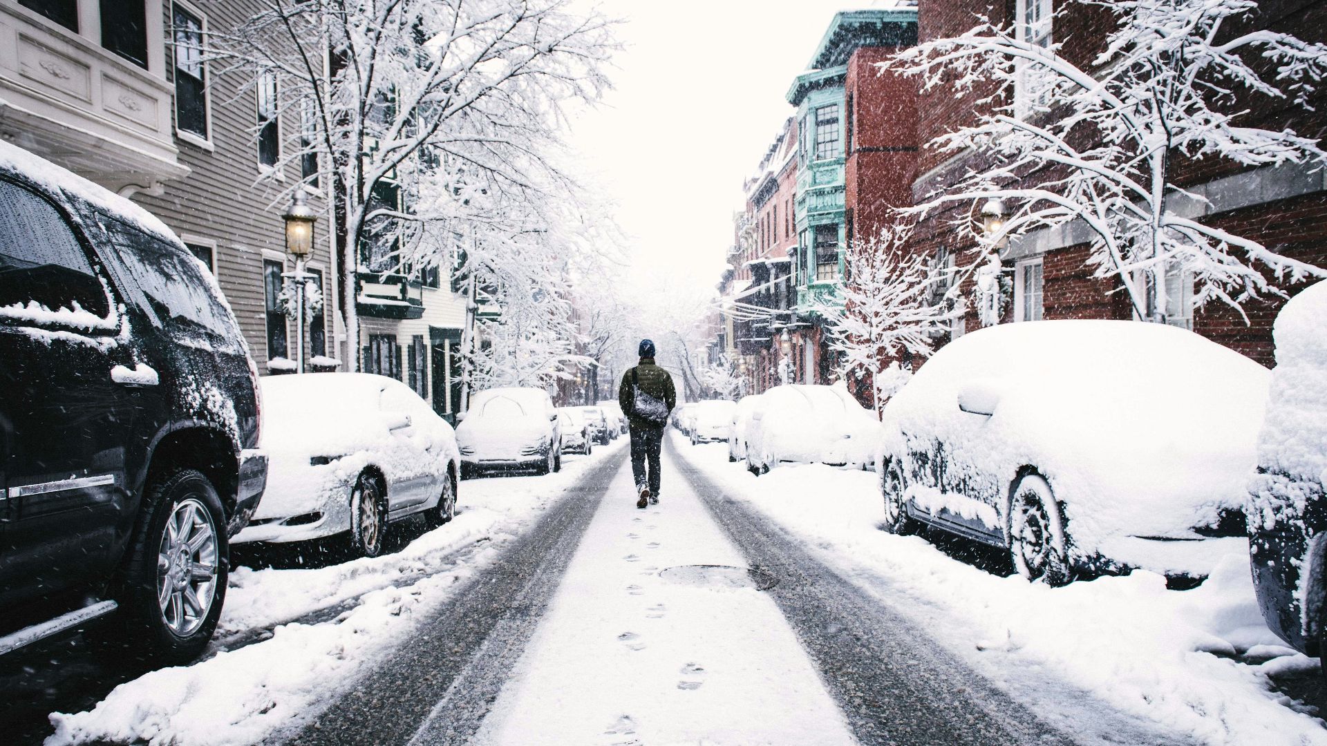 man walking on road covered with snow between vehicles and concrete buildings during daytime