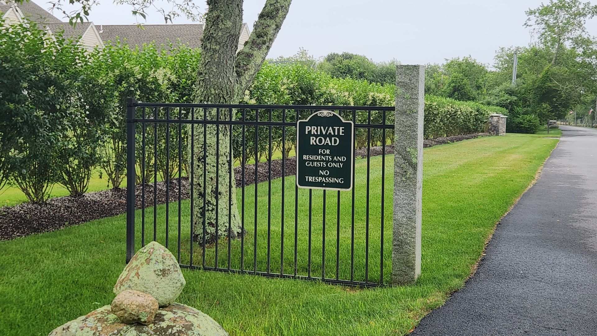 This image shows a private road gate located in a residential area. The gate is a black metal barrier with vertical bars, positioned between two stone pillars. Attached to the gate is a sign that reads