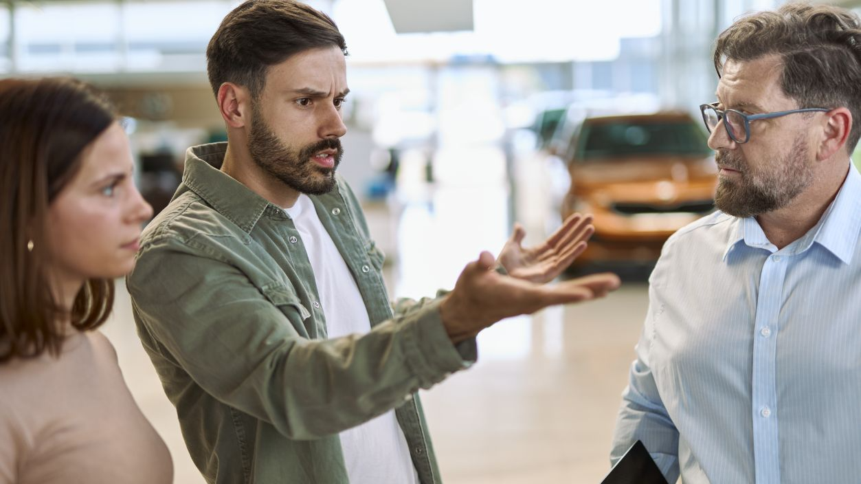 Displeased man and his wife arguing with car salesperson in a car showroom.