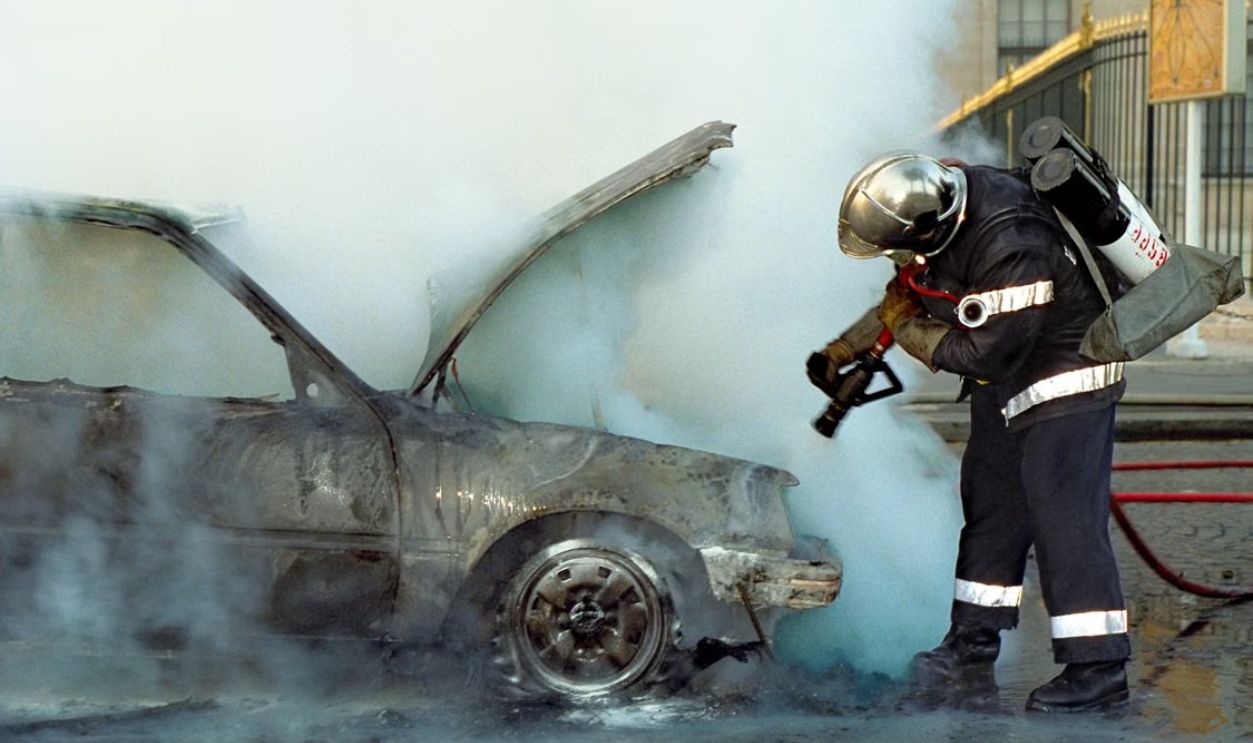 Firefighter using extinguisher on a smoking, damaged car in an urban area. High action scene.