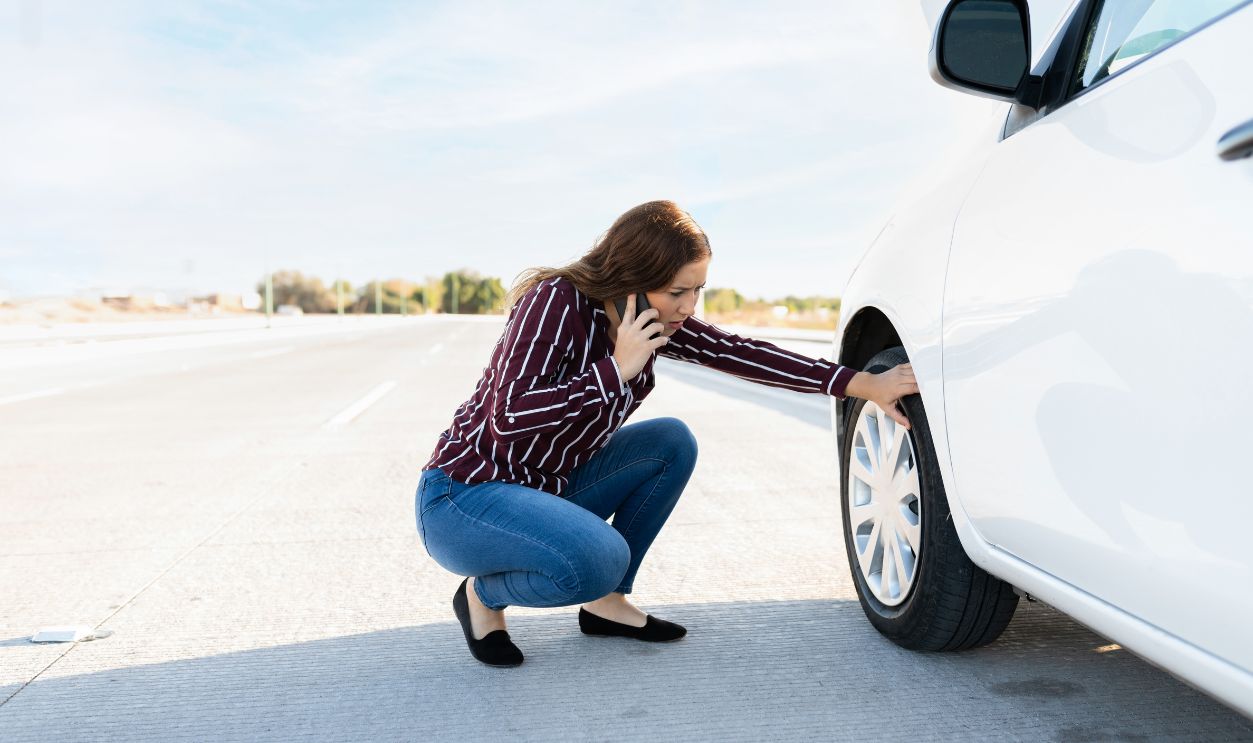 Worried young woman talking on the phone and checking her car flat tire on the side of the highway