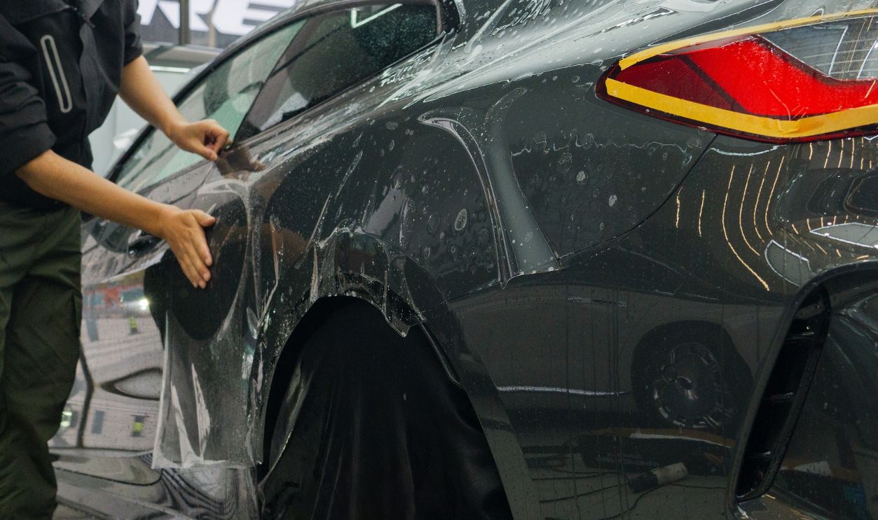 A detailed shot of a skilled technician meticulously installing paint protection film (PPF). Using specialized tools like squeegees and spray bottles, they ensure a flawless, bubble-free application that protects the car's original paint. This image represents premium automotive care, professional craftsmanship, and investment in vehicle preservation, ideal for marketing luxury detailing services, PPF installers, or illustrating top-tier automotive aftermarket services.