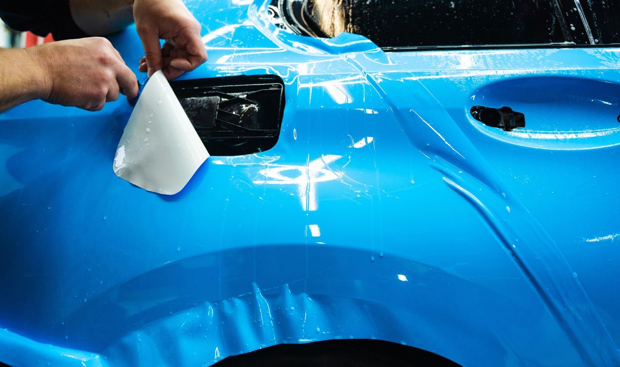 Detailing technician trims blue PPF on a car panel for perfect contour fit. Cutting blue PPF with blade. Cutting blue paint protection film with blade during detailing job ensures precision edges
