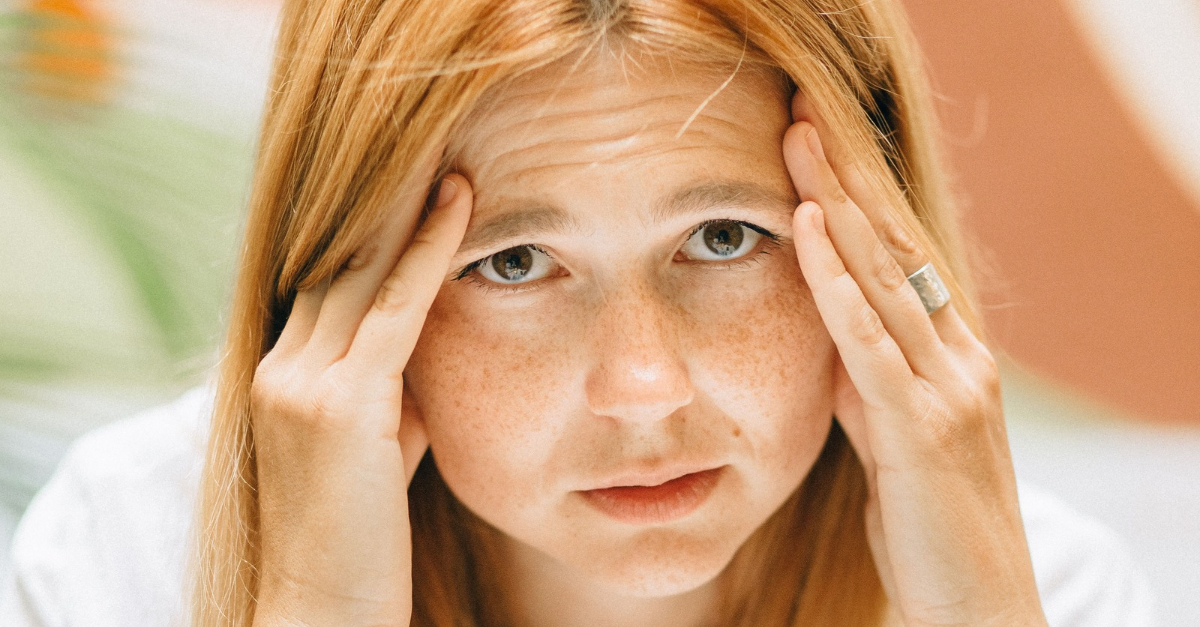 Close-up of a woman sitting at a desk with hands on her head, looking stressed and exhausted.