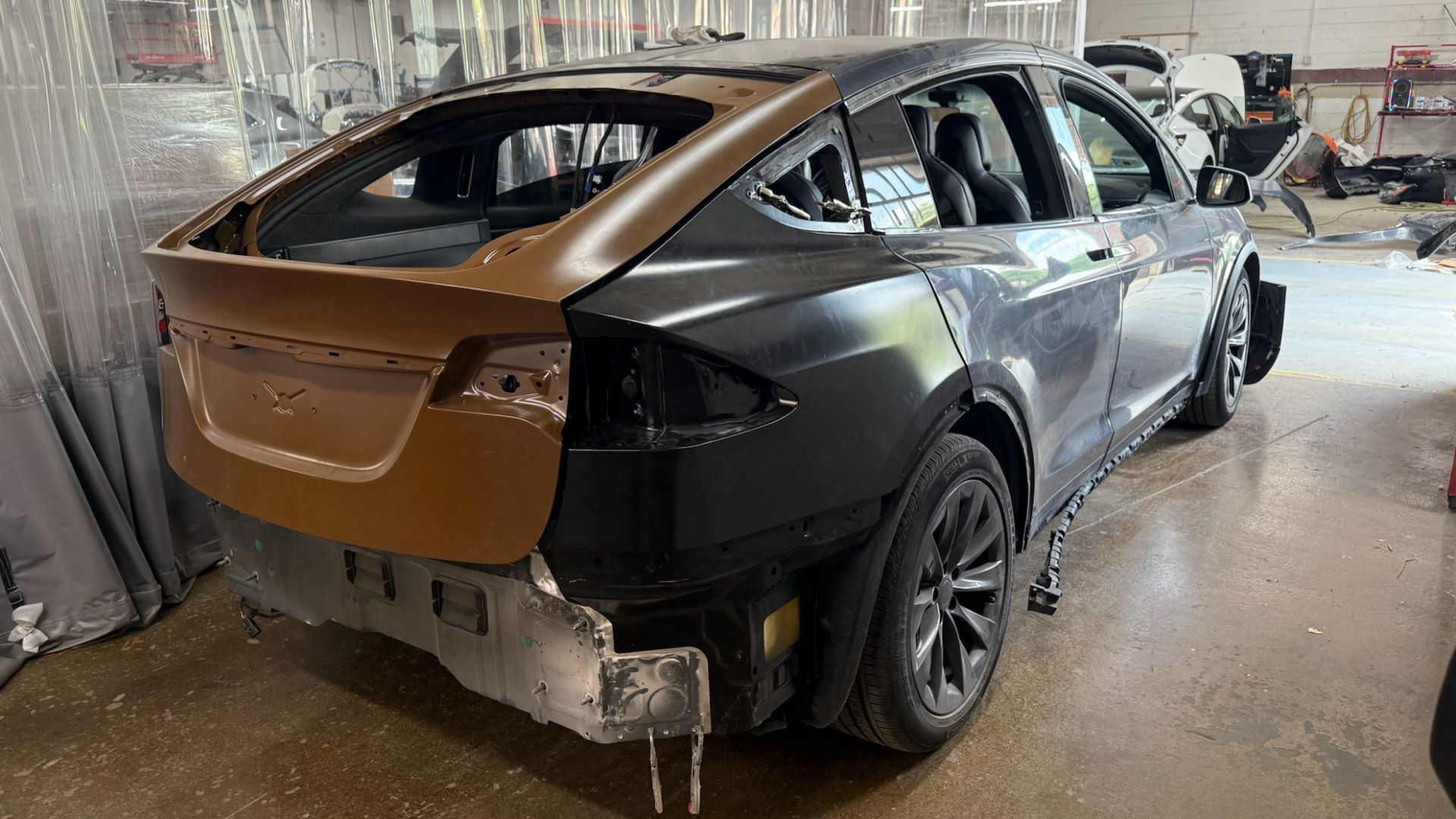 This image shows the rear section of a Tesla Model X undergoing collision repair inside an auto body shop. The liftgate and rear panels have been removed or replaced with unfinished parts, exposing portions of the vehicle’s frame and structural components