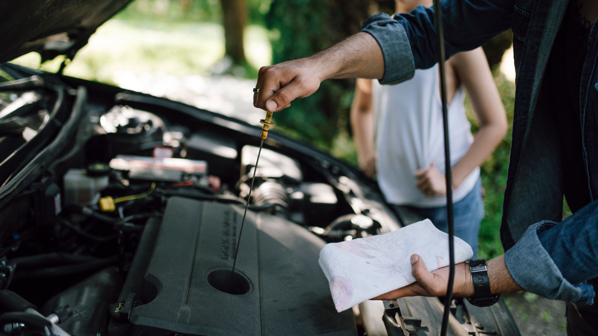 Checking oil level in a car engine during a sunny afternoon with a child observing nearby