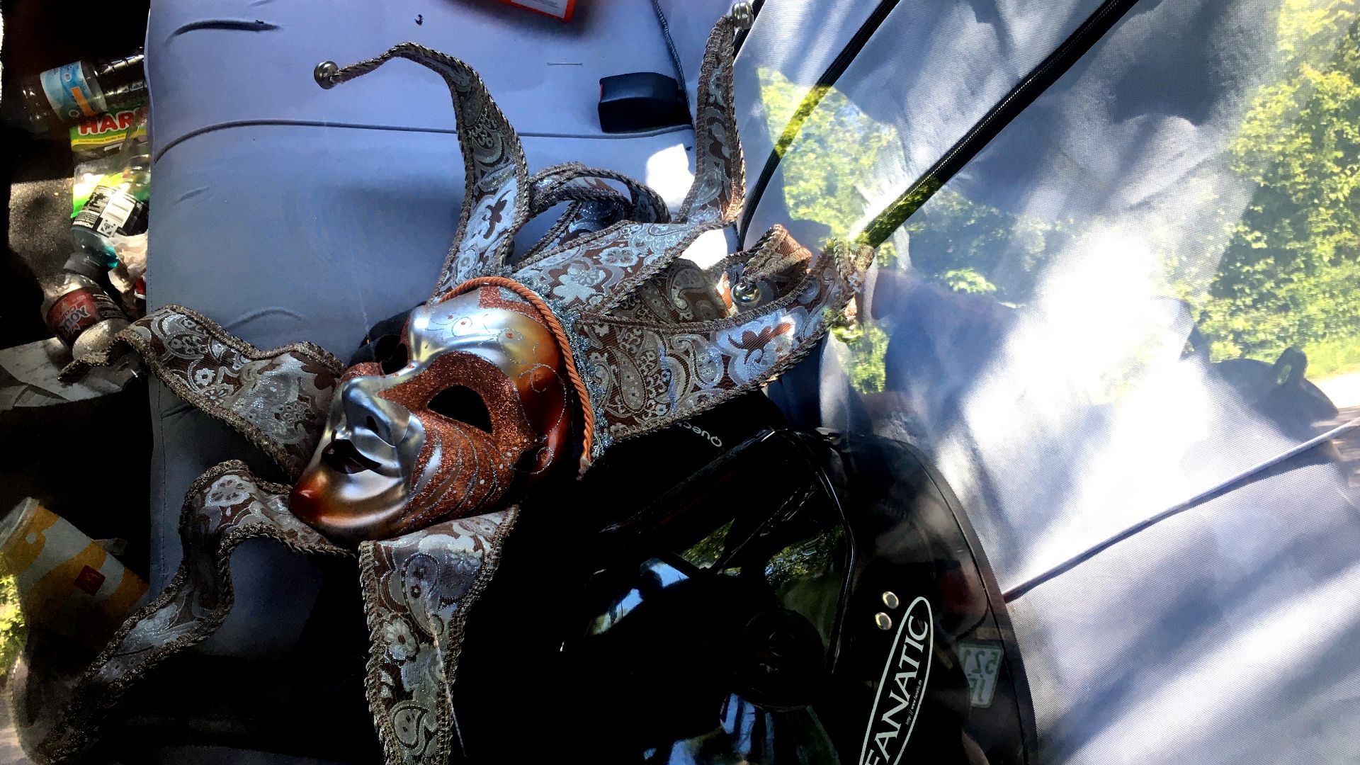 A Carnival-in-Venice-mask on the backseat of a parked car, together with various items like a Amazon Fire TV stick, a motorcycle helmet, a McDonalds softdrink container, an empty fruit gum plastic bag, and the like. Further, there are reflections of the p
