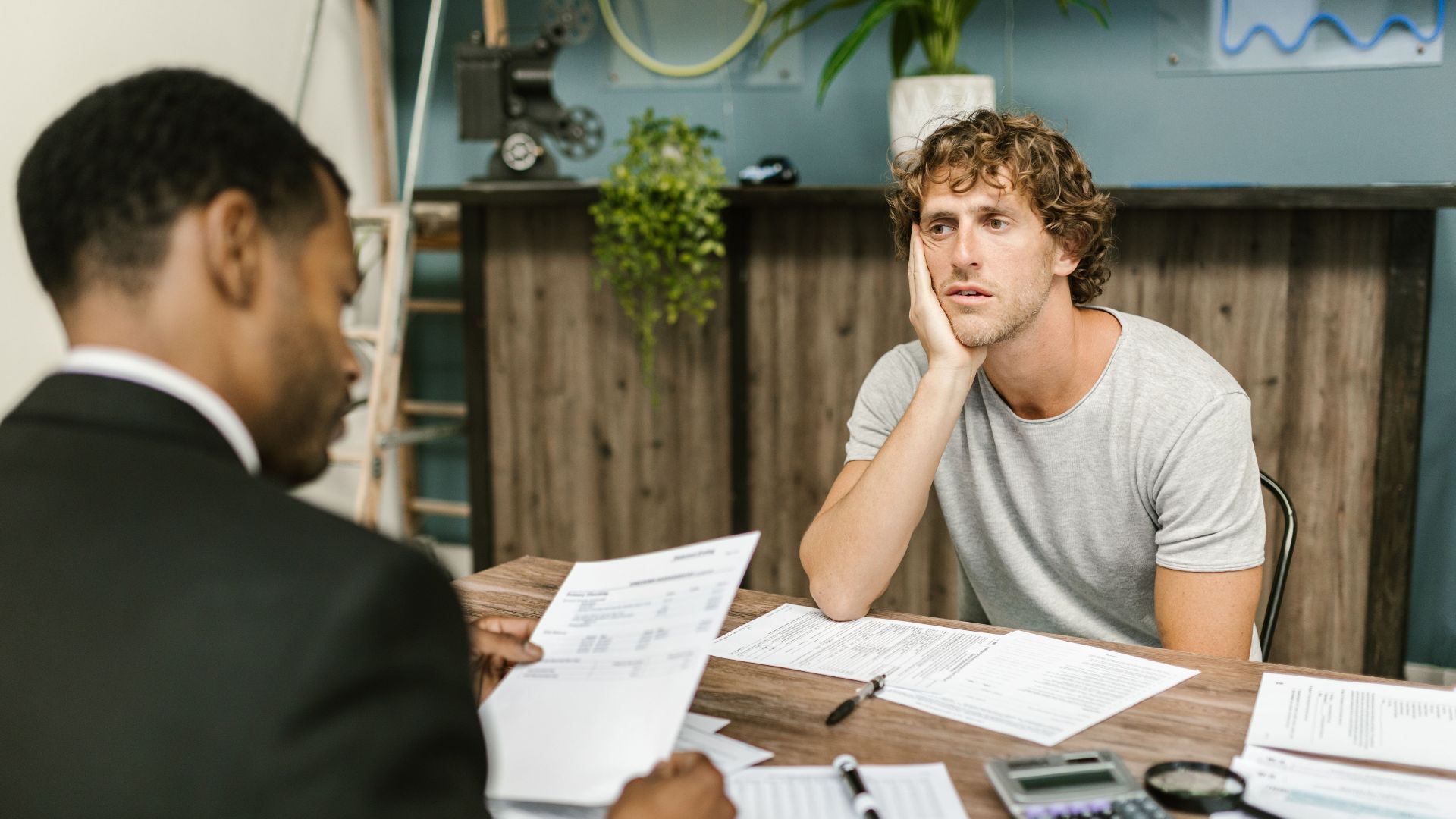Man in office, appearing stressed during paperwork discussion, sitting opposite professional.