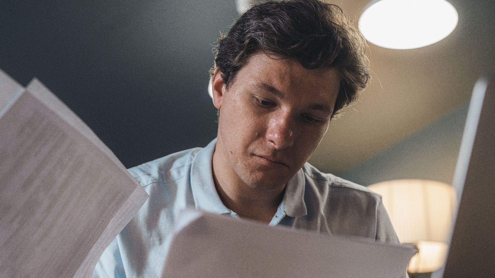 Serious man wearing a light blue shirt intently reading printed papers indoors under warm lighting.