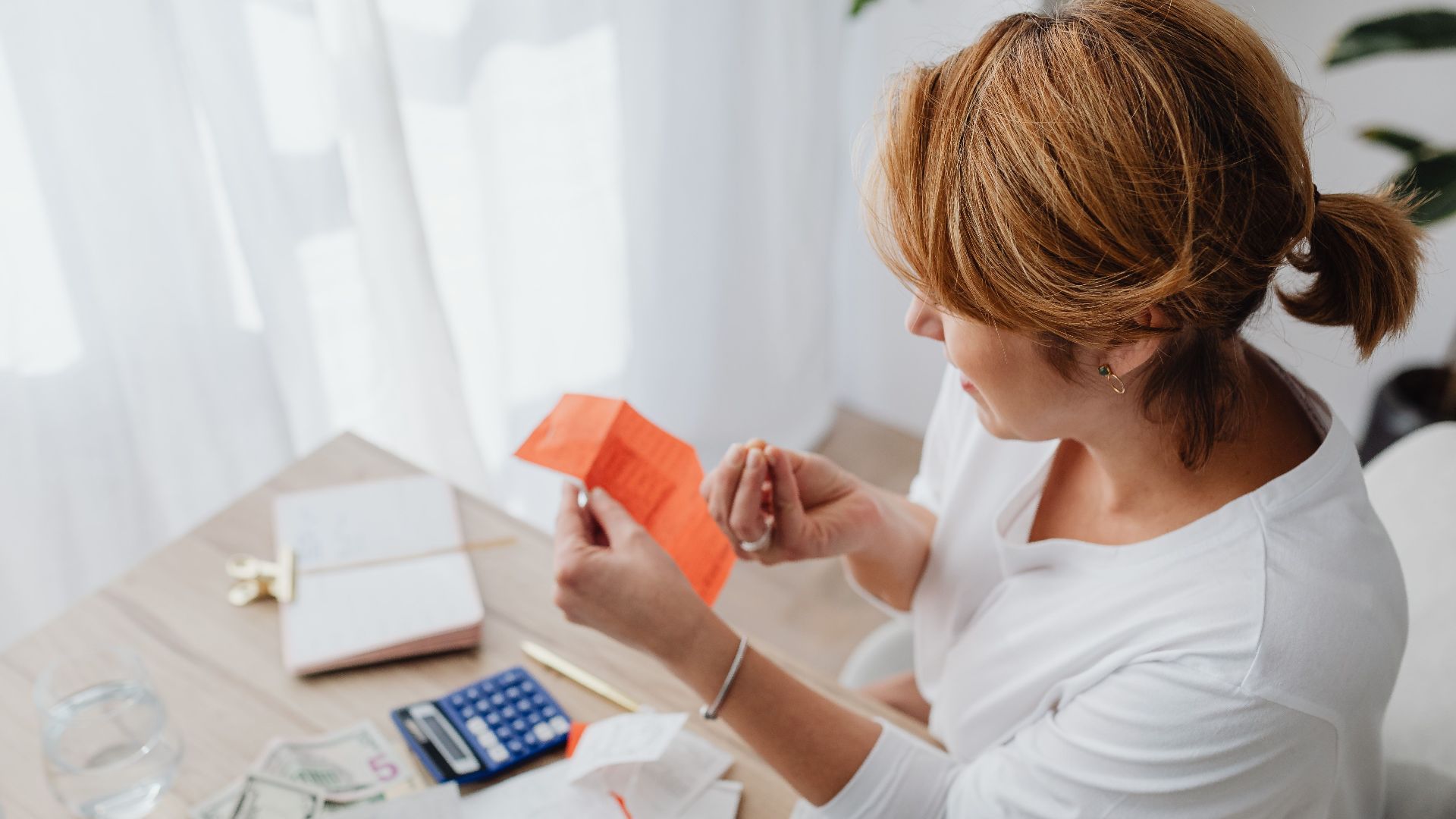 A woman organizing her finances with bills and receipts at a home office desk using a calculator and laptop.