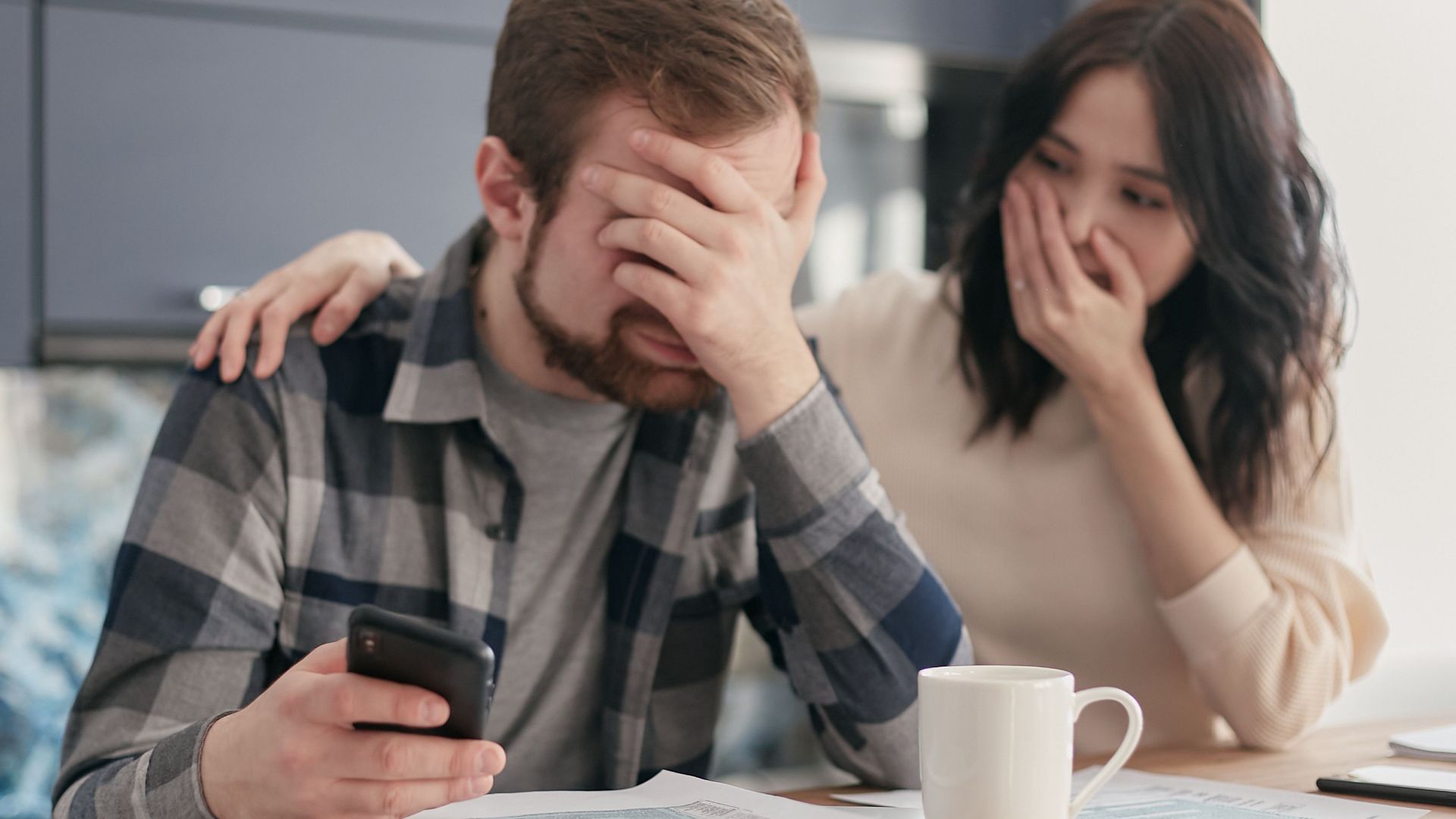 A couple looking stressed and holding a cellphone while reviewing financial documents indoors. Hands on faces in concern.