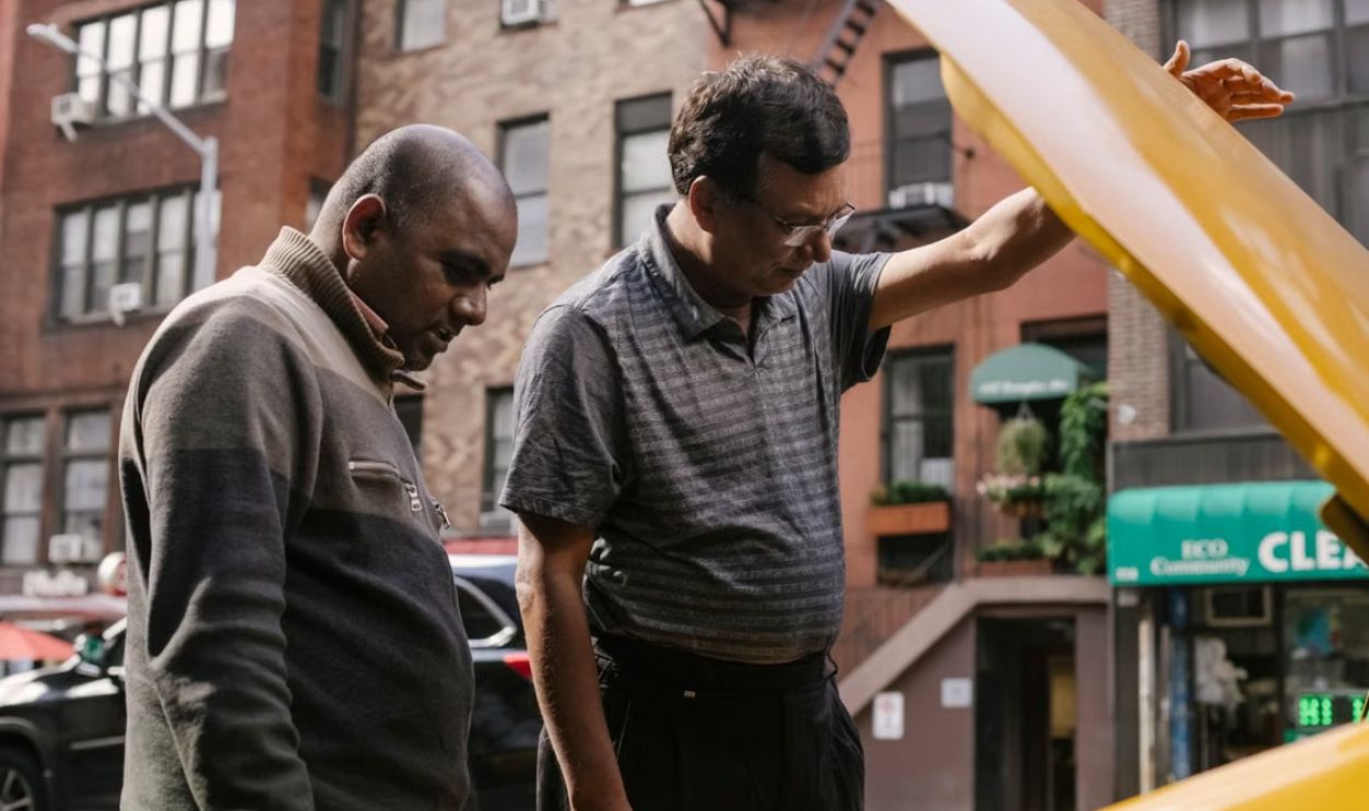 Two men check a car engine on a city street, problem-solving in an urban setting.