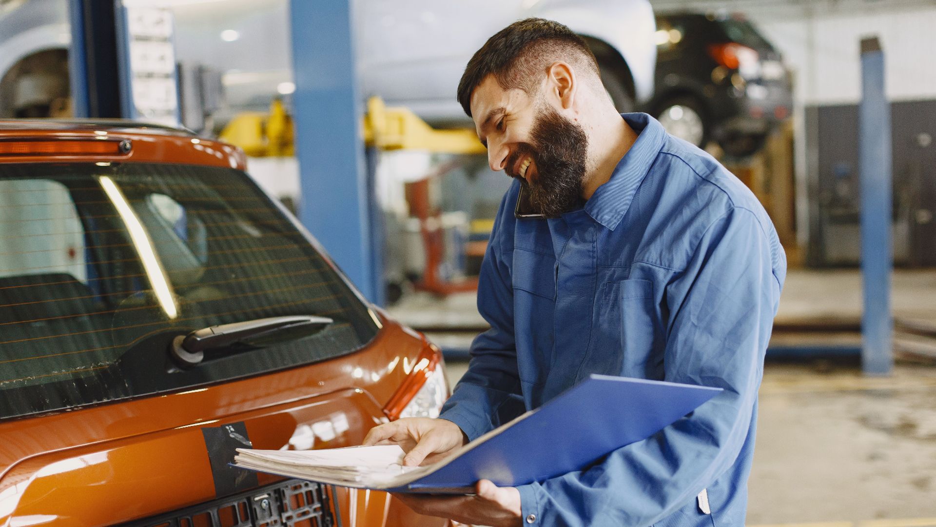 Smiling auto mechanic inspects a car in a service garage, wearing coveralls and holding documents.