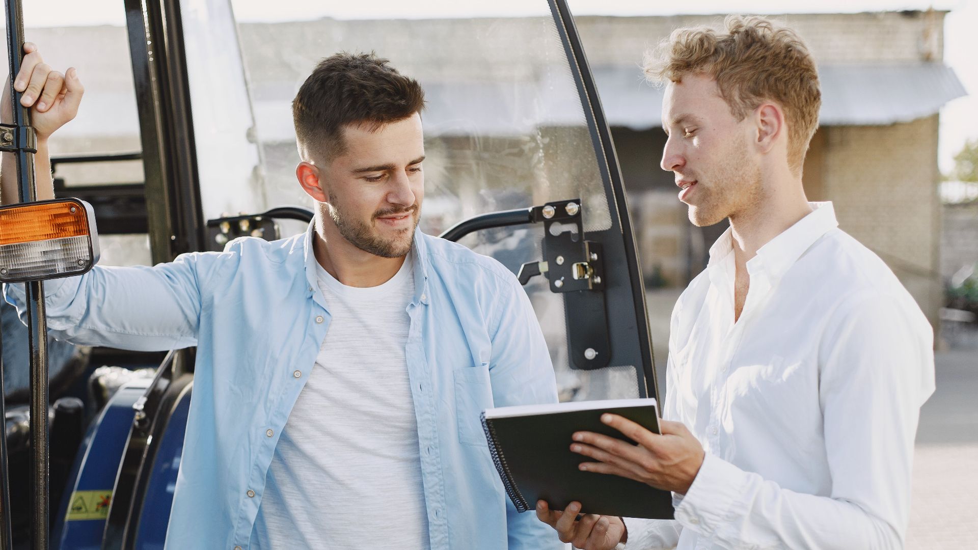 Two men discussing business plans beside a blue tractor outdoors.