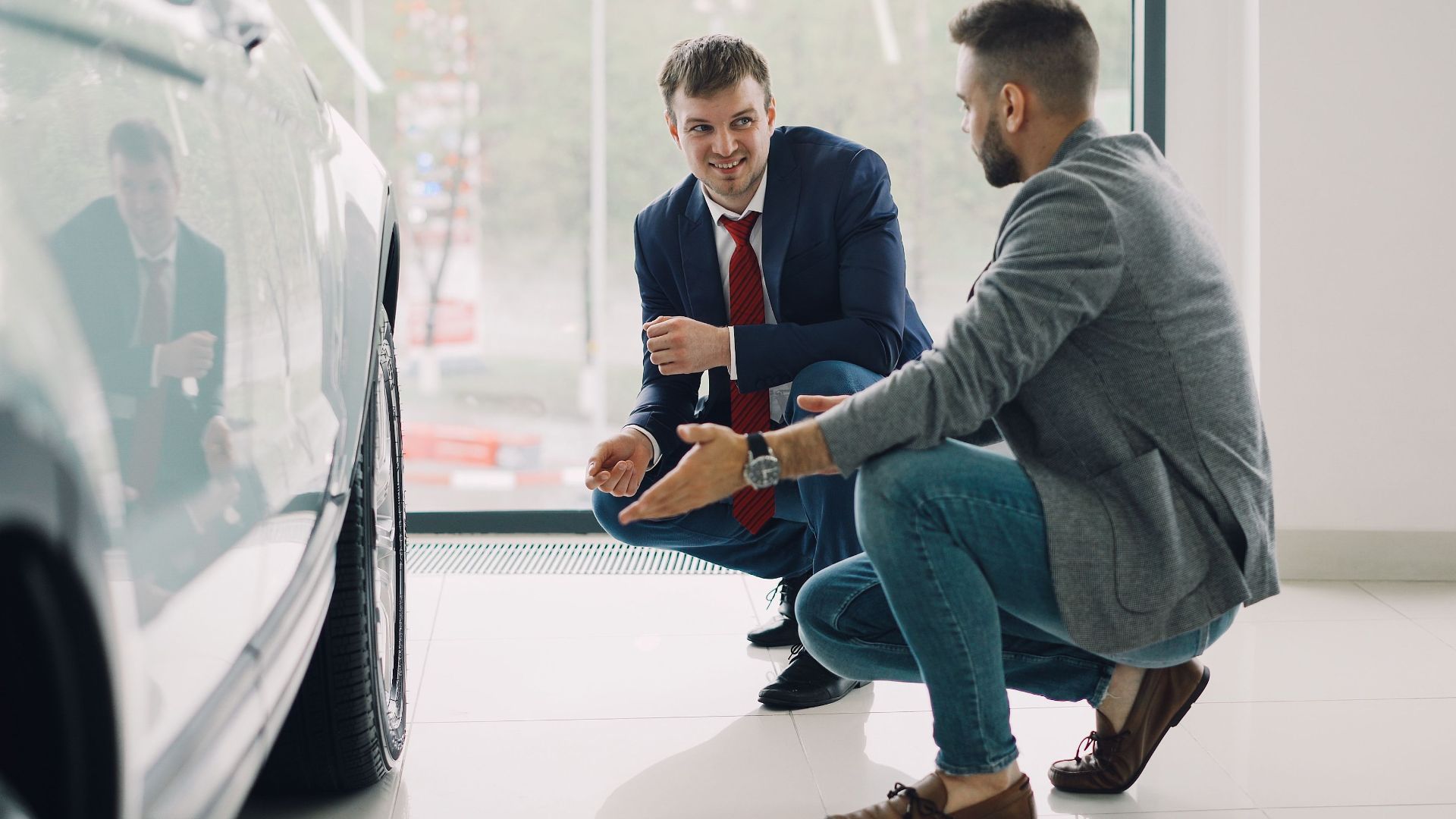 Two men discussing car features in a showroom, kneeling near a vehicle.