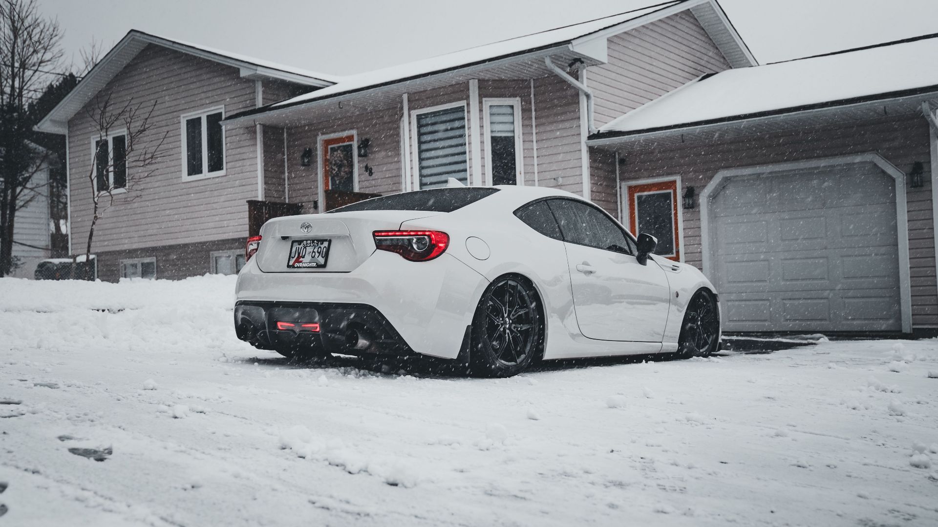 A white sports car parked in a snowy suburban driveway during winter snowfall.