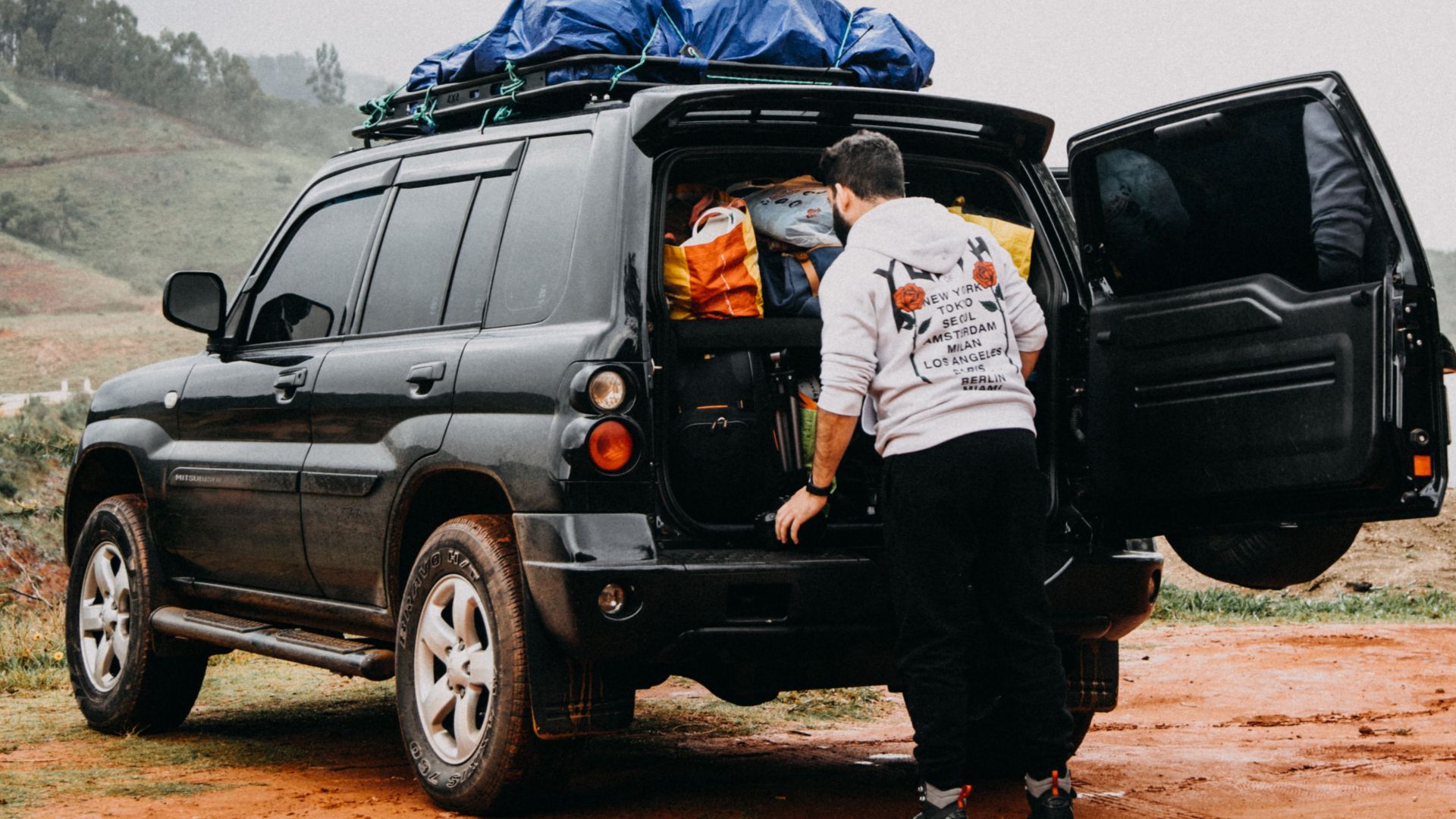 A man packs an SUV with luggage on a dirt road, preparing for an outdoor adventure.