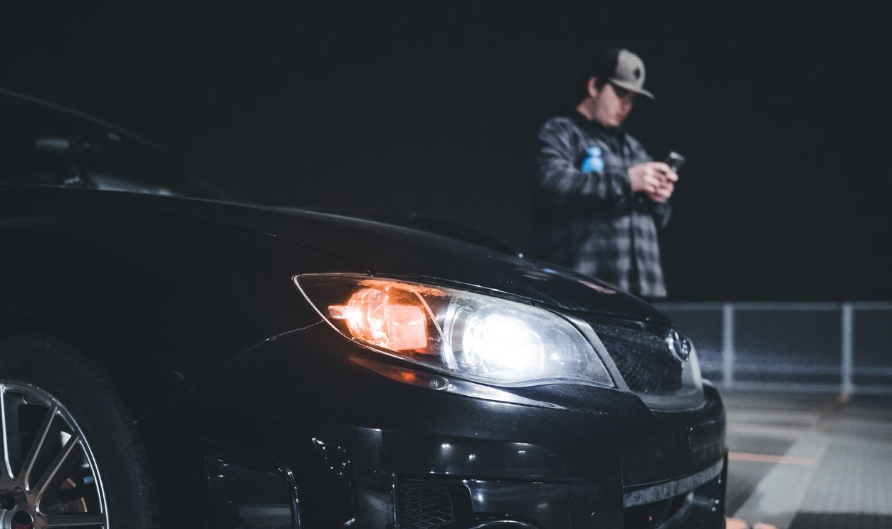 A man stands near a black car at night, using his smartphone in an urban setting.