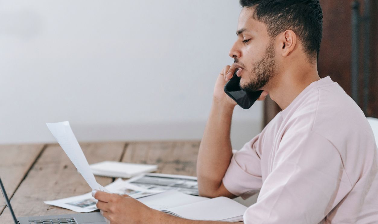 A young man conversing on a smartphone while working from home, reviewing documents and using a laptop.