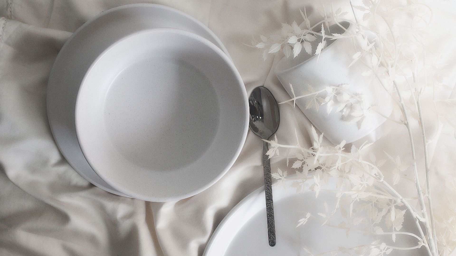Top view of a minimalist table setting with white ceramics, stainless steel teaspoon, and leaf decor.
