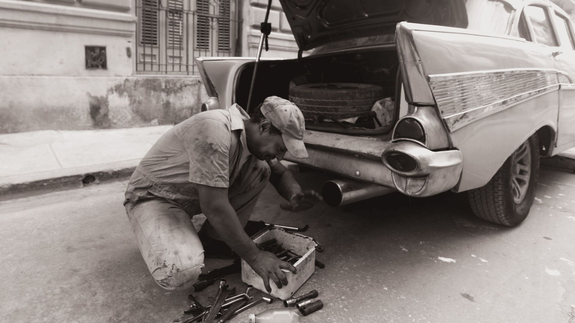 A mechanic repairs a vintage car on the street, surrounded by tools.