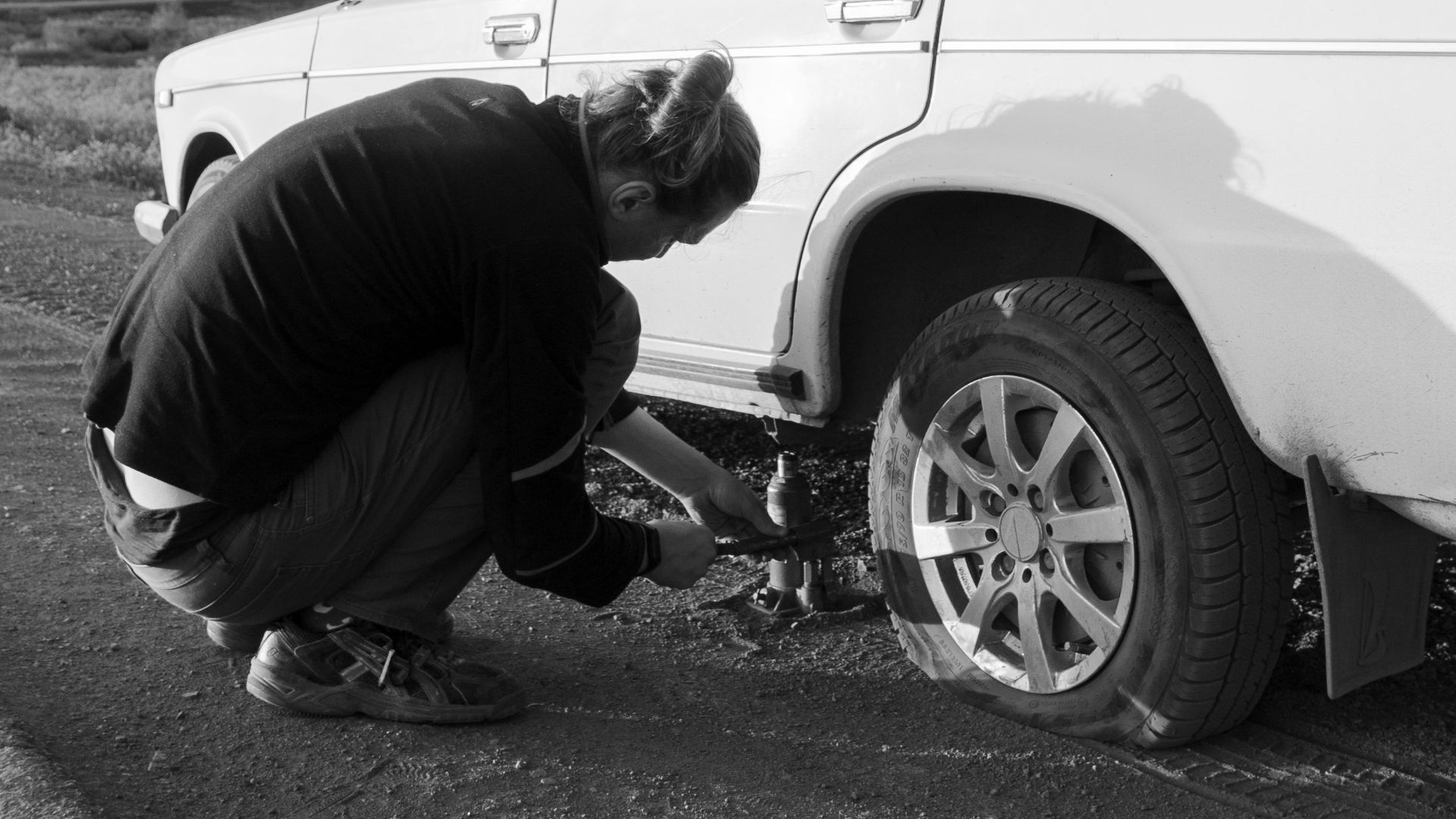 A man changes a car tire on a country road. Black and white photo.
