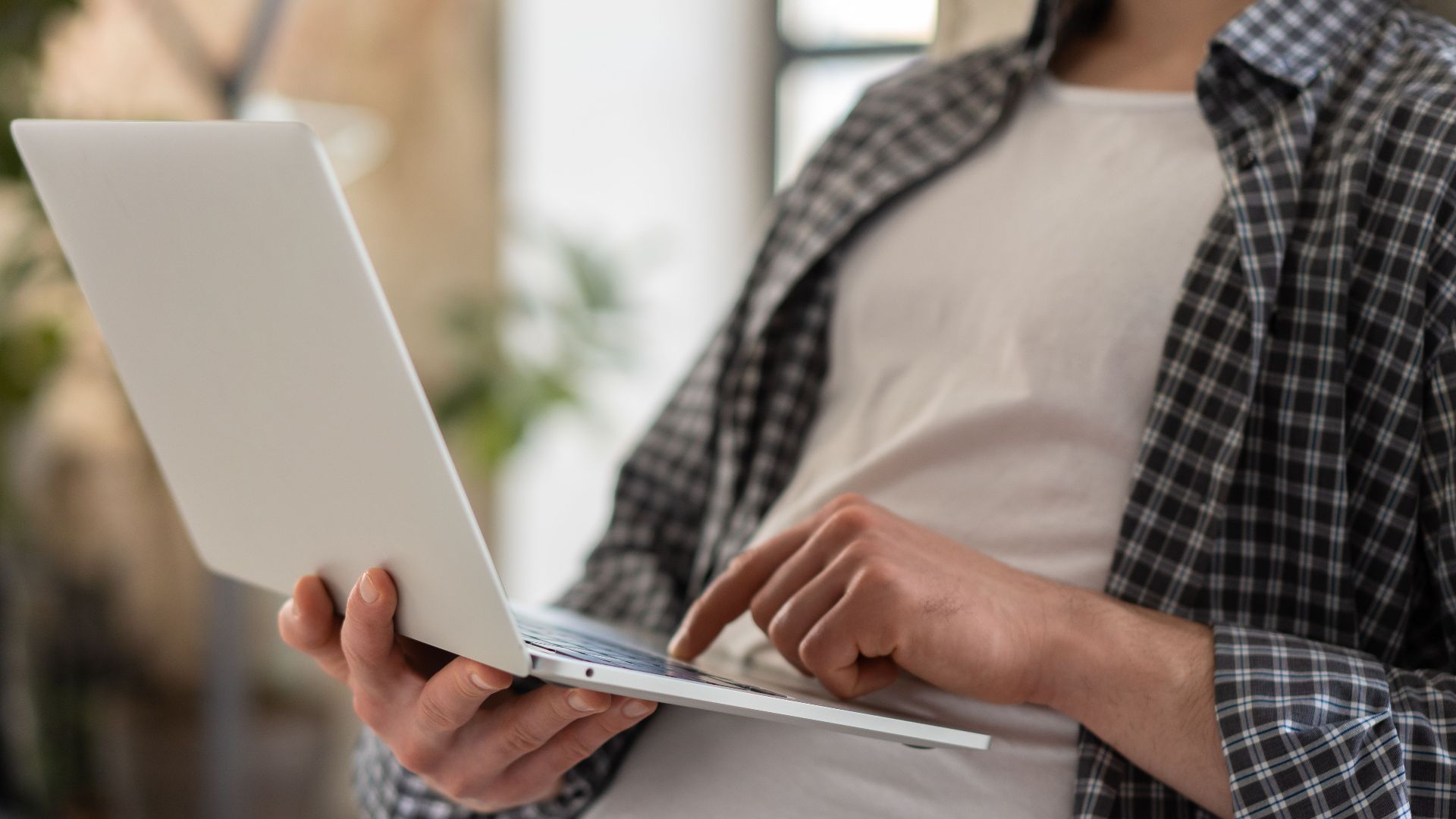 Young adult in casual shirt using a laptop indoors with blurred background and shallow focus.