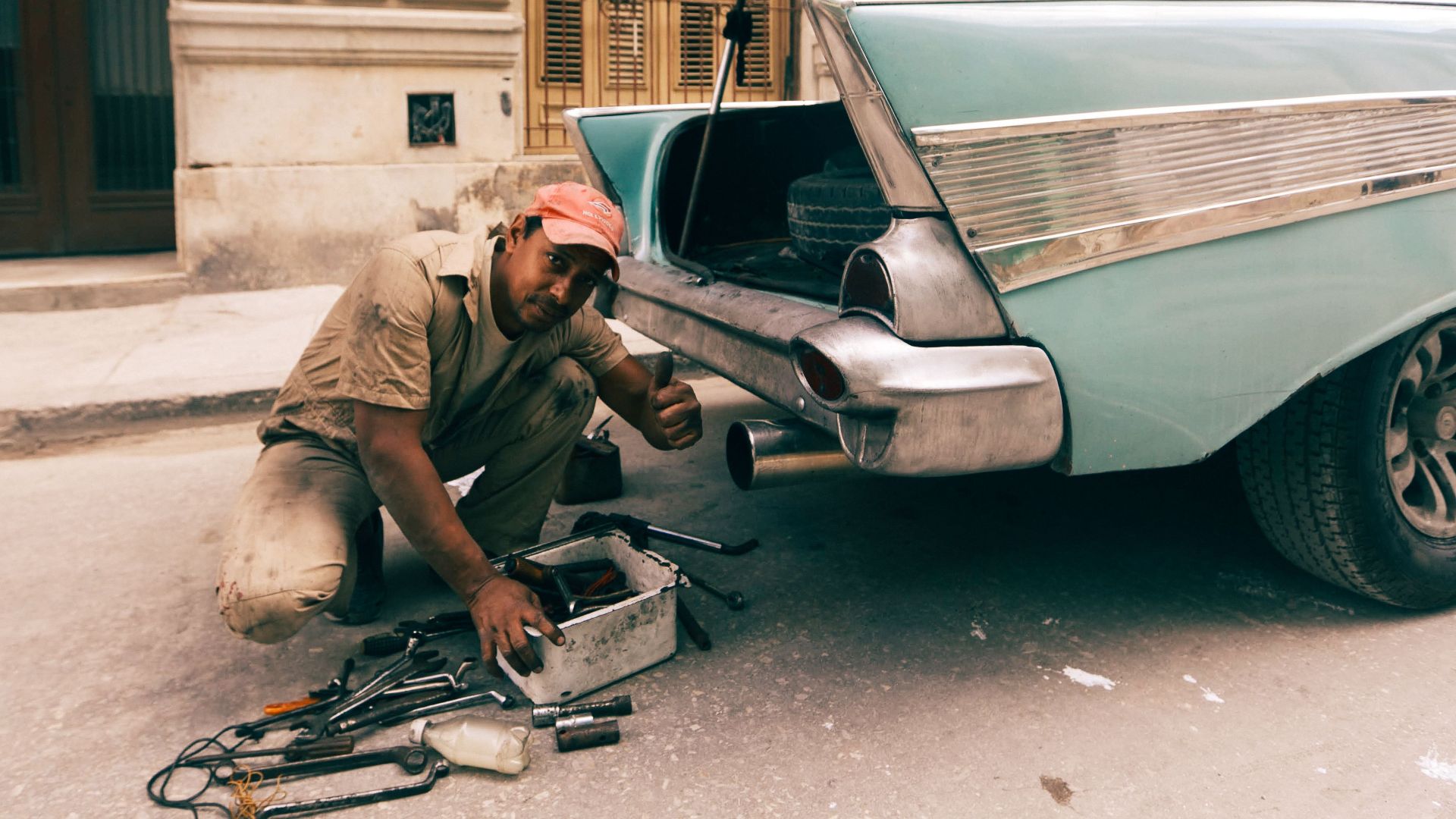 Mechanic gives thumbs up while working on classic car trunk outdoors.