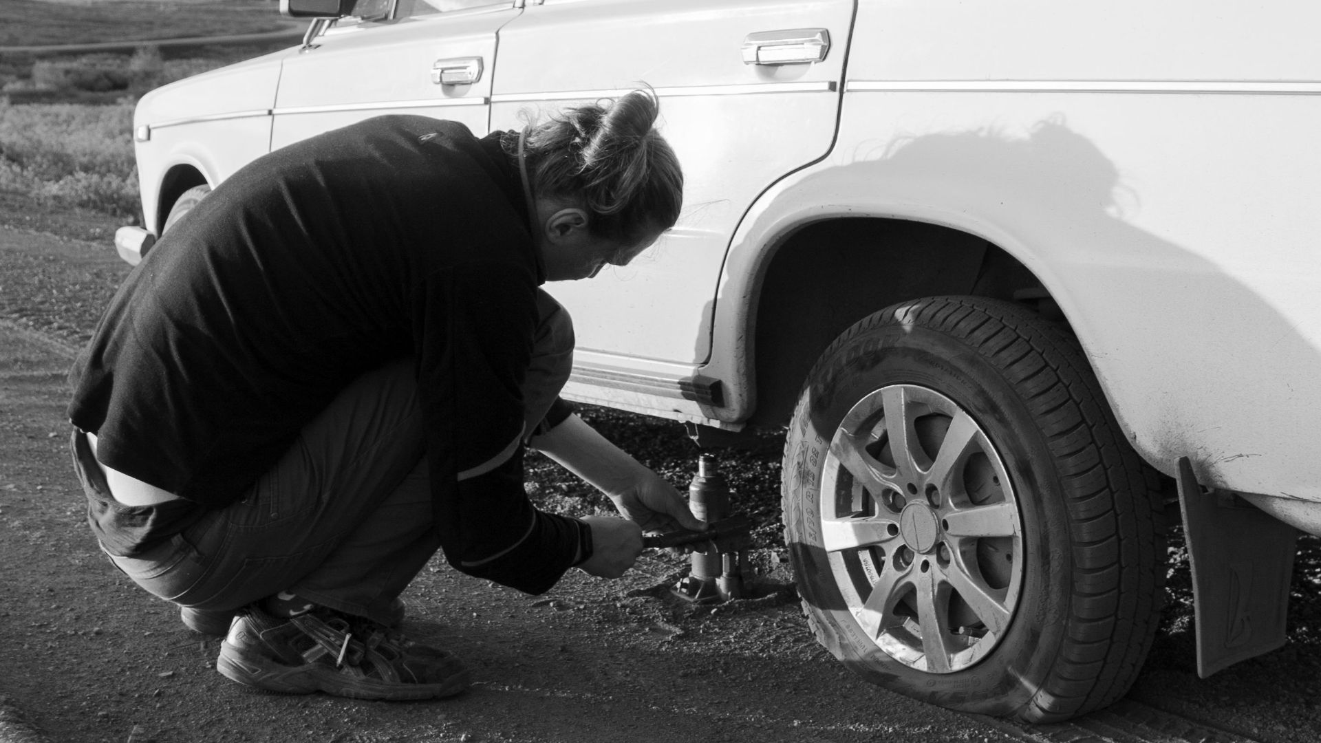 A man changes a car tire on a country road. Black and white photo.