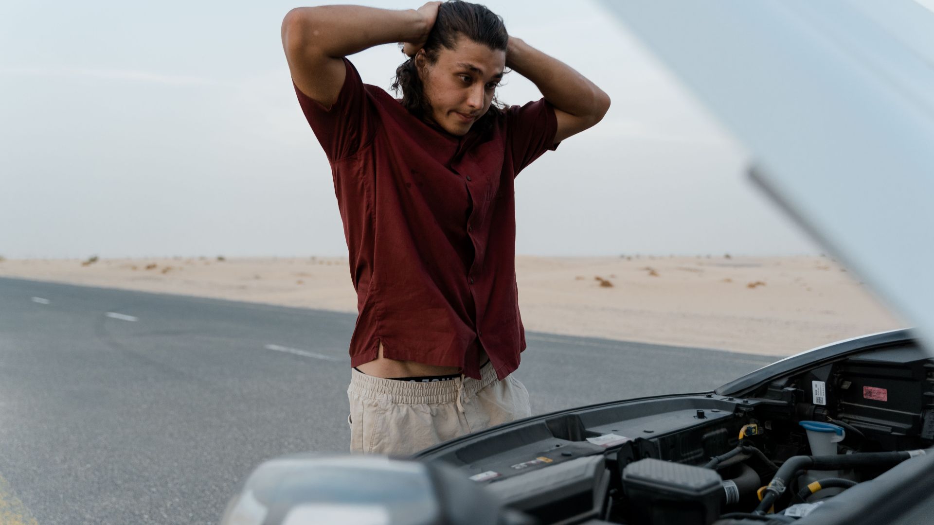 A frustrated man examining a broken car engine on a deserted road, showing stress and concern.