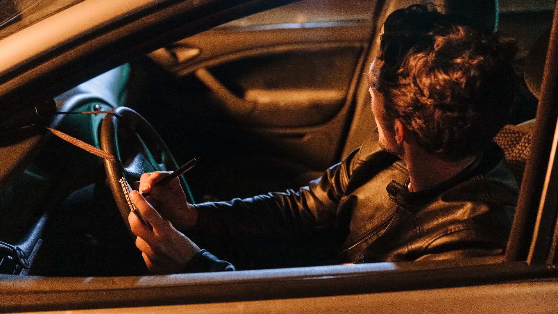 A man in a leather jacket taking notes inside a car at night, implying a stakeout or investigation setting.