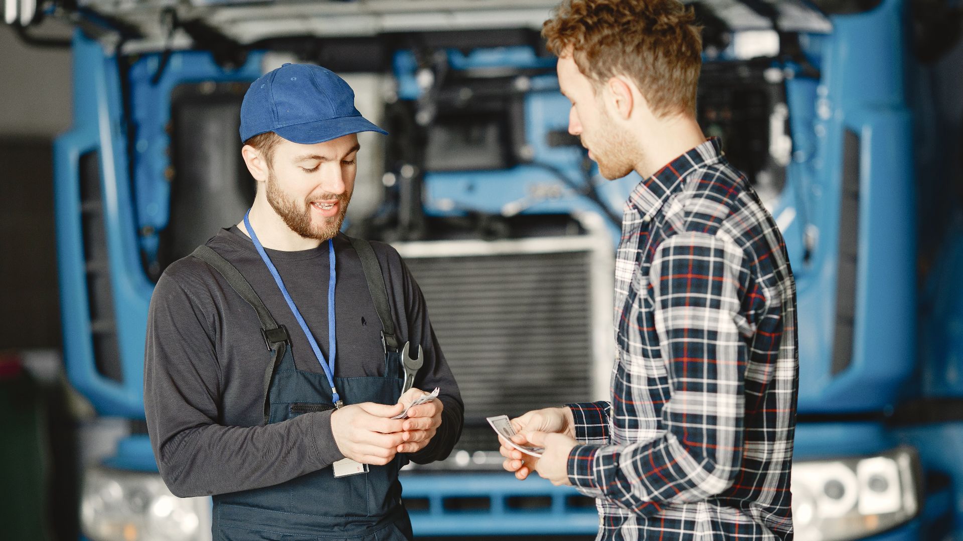 Auto mechanic in blue uniform receives payment from customer in a garage setting with a truck in the background.