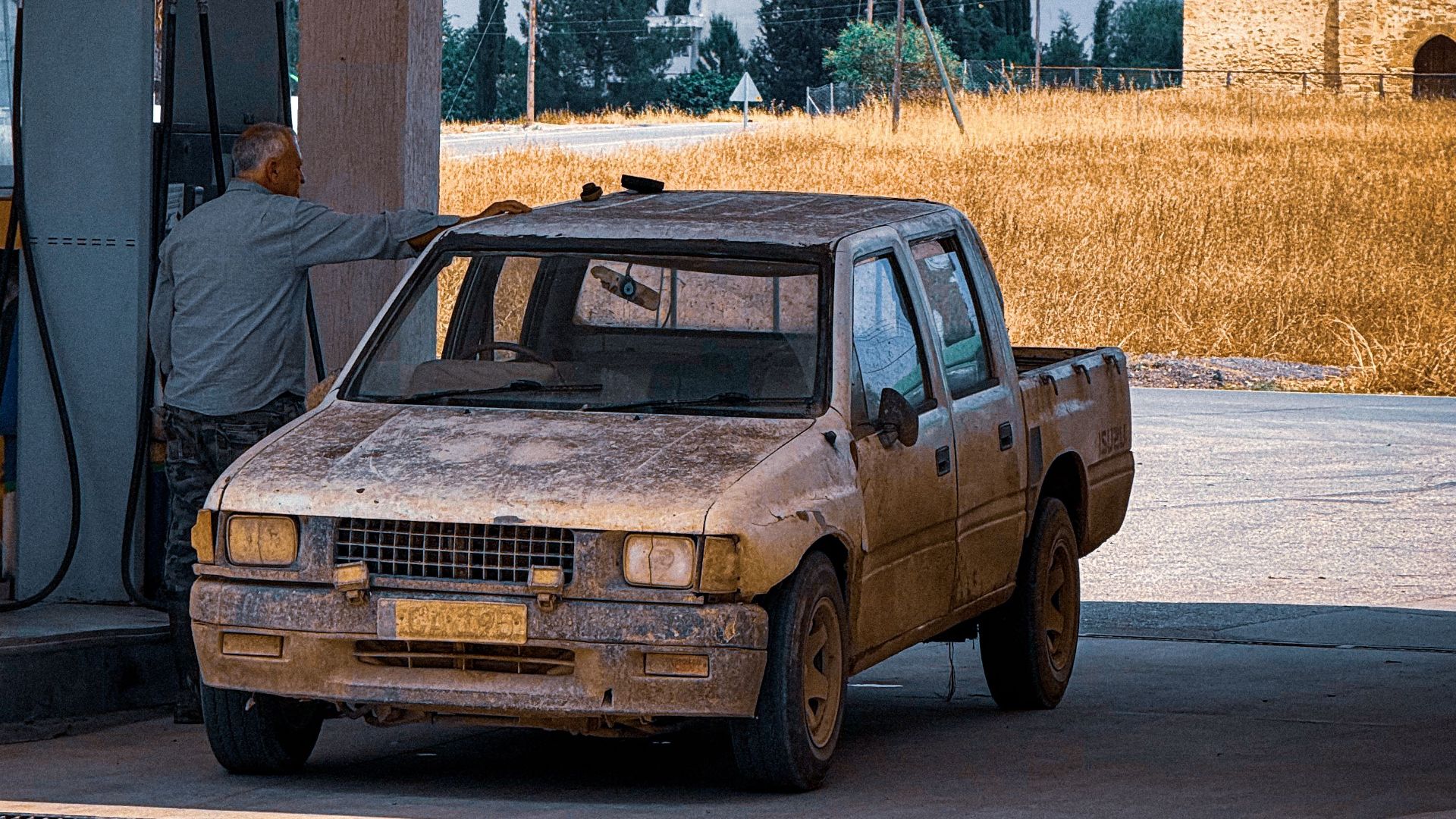 A weathered truck refueling at a gas station against a rural Cypriot backdrop.