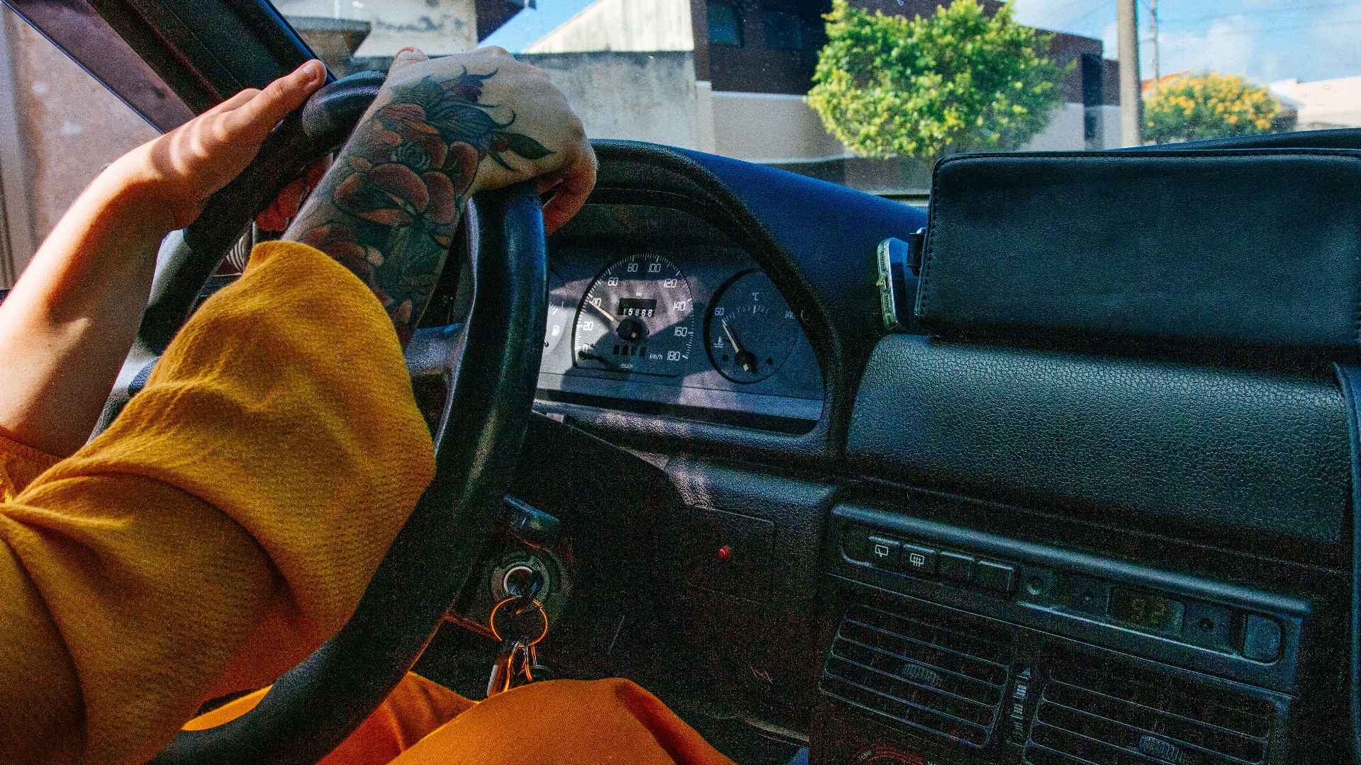 Close-up of a tattooed person driving a vintage car with a colorful dashboard.