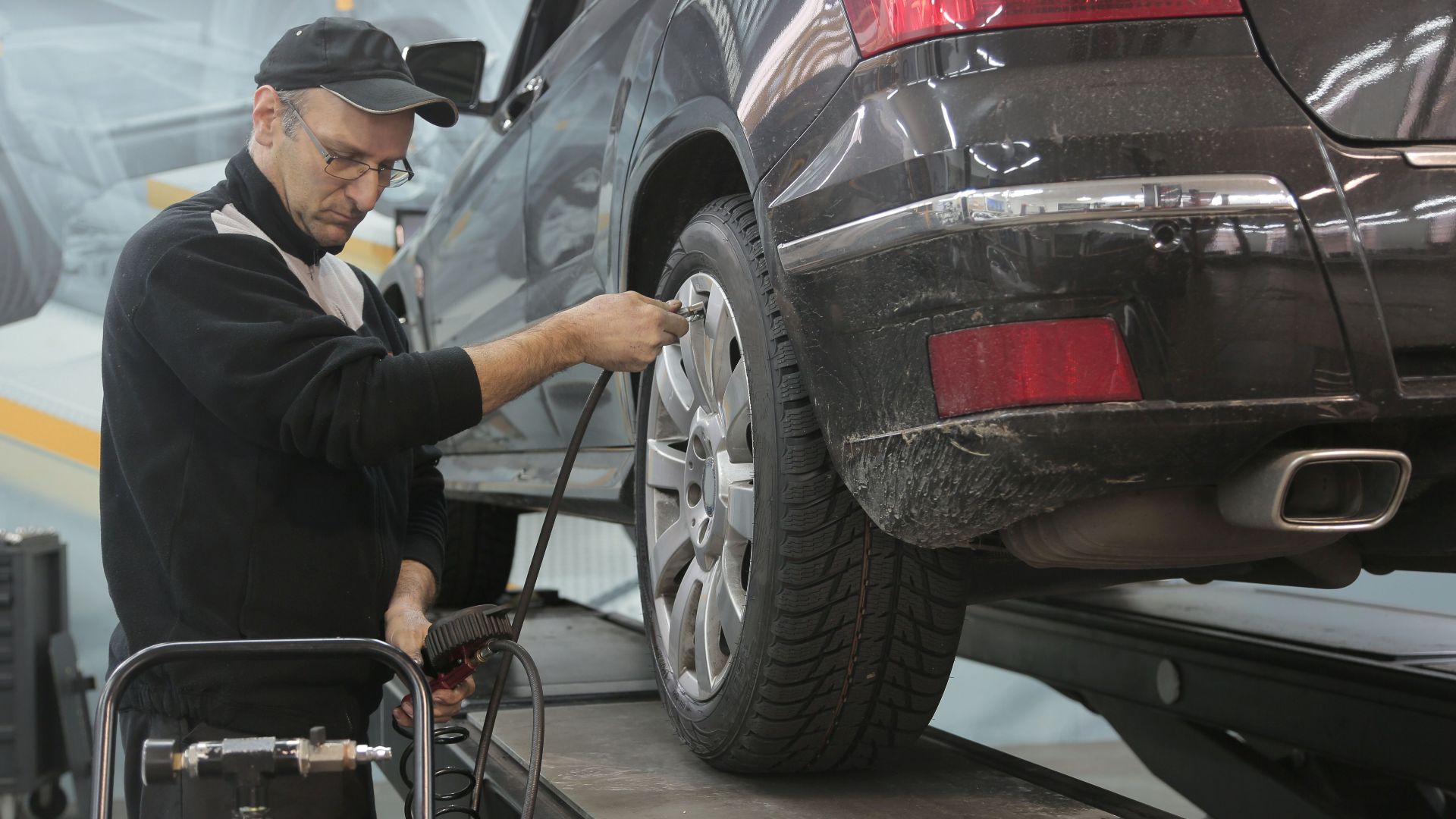 Side view of professional serious auto mechanic in casual uniform and eyeglasses pumping up car wheel while working in modern car service