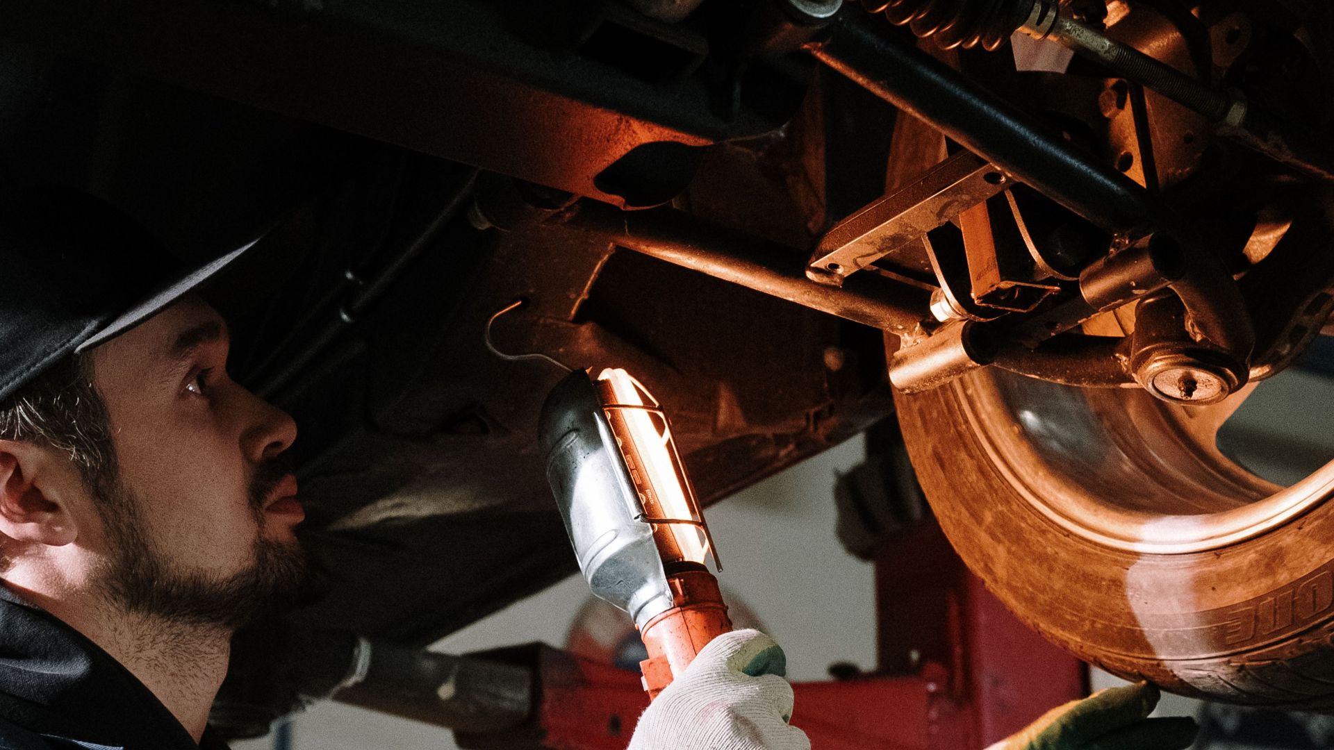 Mechanic uses a flashlight to inspect car's suspension in a garage setting.